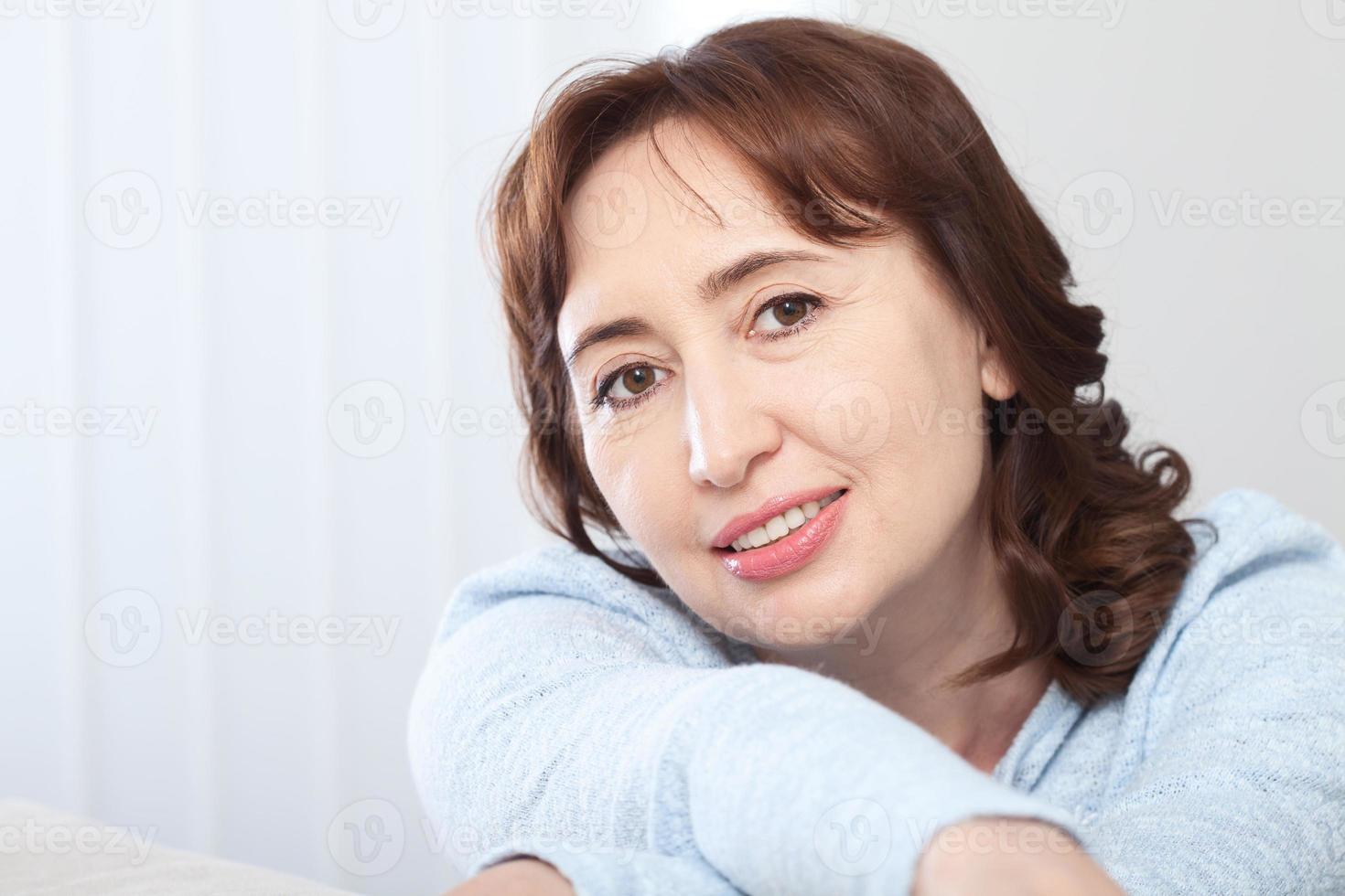 Lovely middle-aged brunette woman with a beaming smile sitting on a sofa at home looking at the camera photo