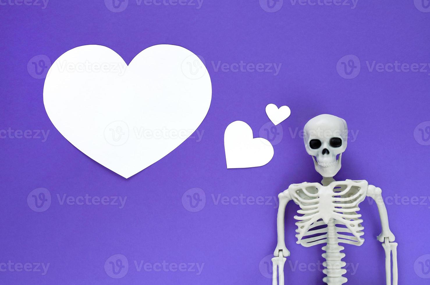 Skeleton on violet background with white blank paper cut hearts, one big heart with copy space. Anatomical plastic model human skeleton with dreaming text balloon. Valentine's day love concept. photo