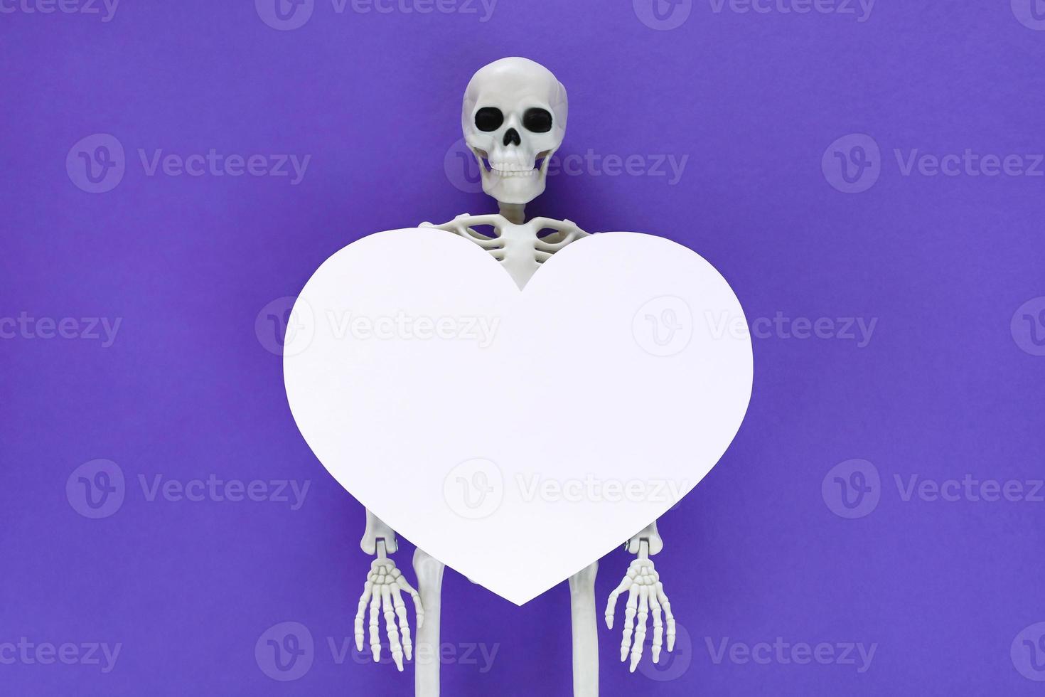 Skeleton with big white blank cut out heart on it, place for text in paper heart frame. Anatomical plastic model human skeleton centered on violet background. Valentine's day love concept. photo