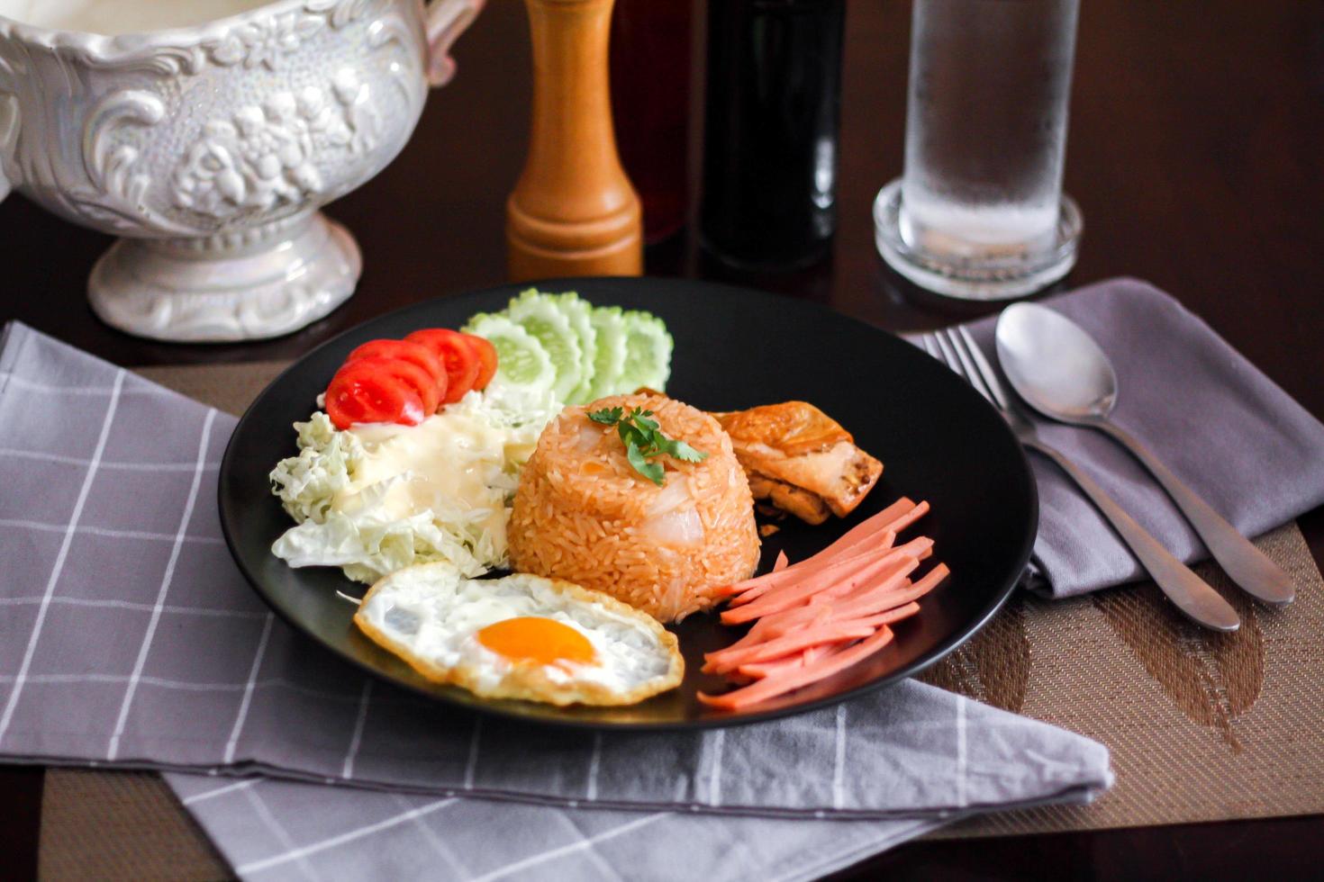 Fried rice, fried egg, cucumber, sliced tomato, green salad and Fried chicken placed on a black plate as a garnish. photo