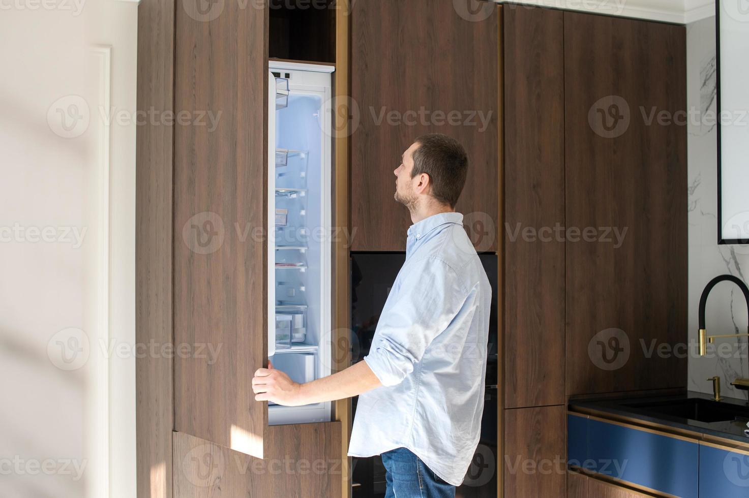 A man stands near an open empty refrigerator and studies the contents photo