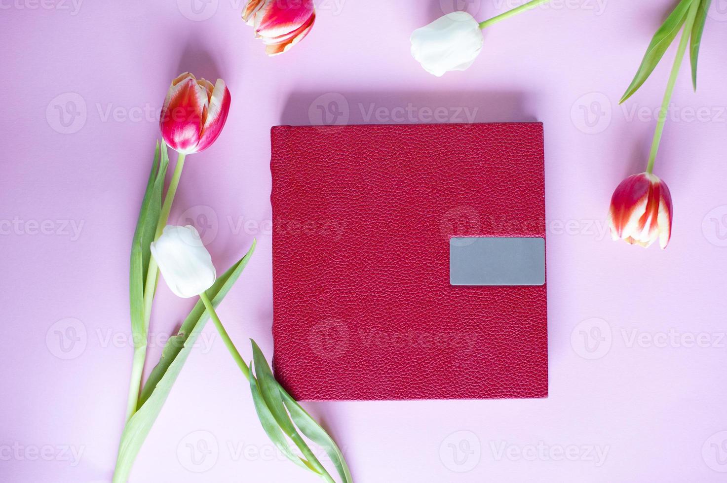 A red leather book with an embossed place for text, lies on a purple background next to flowers and tulips photo
