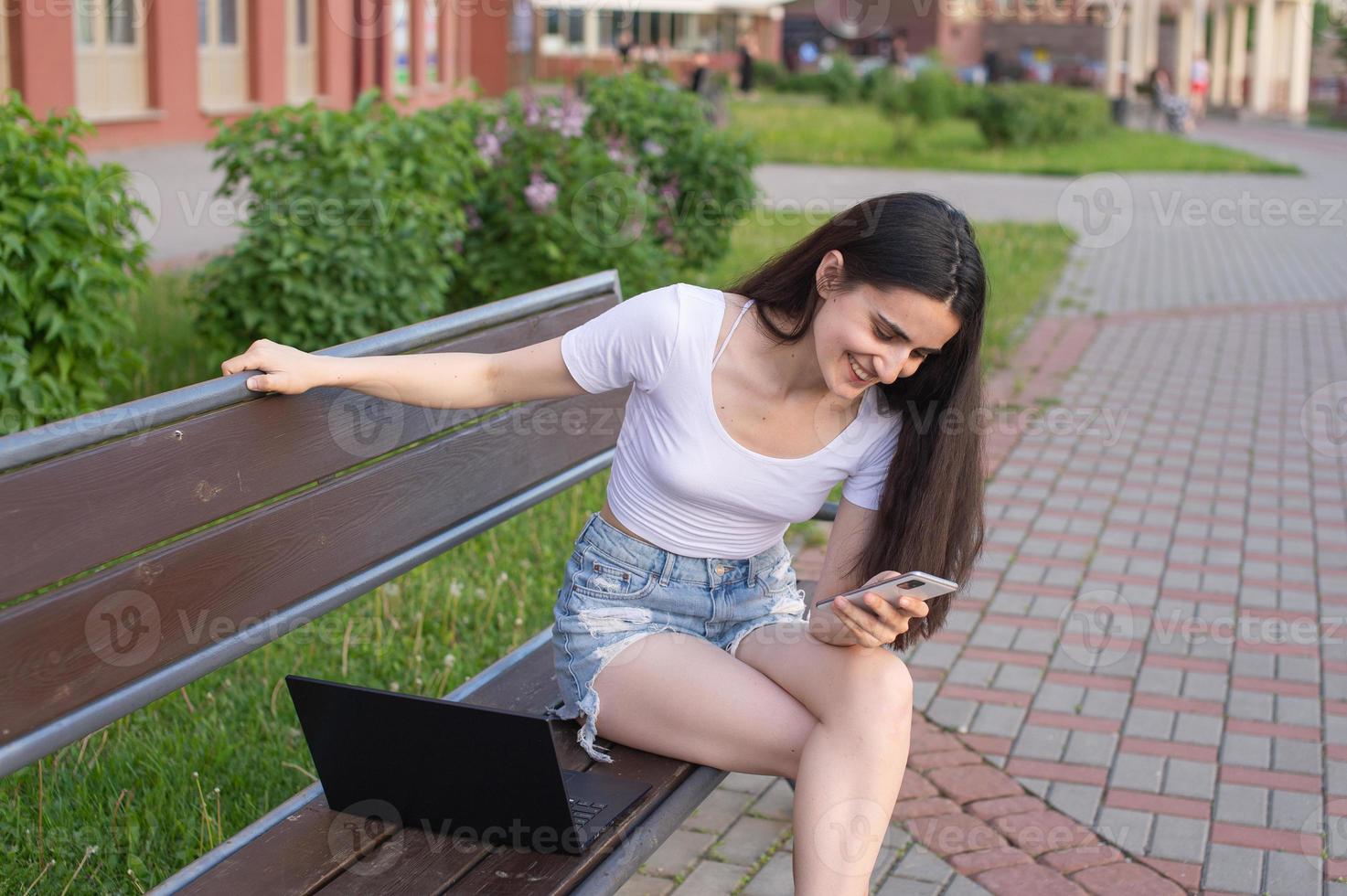 The girl is sitting on a bench, laughing and looking at the phone, there is a laptop nearby photo