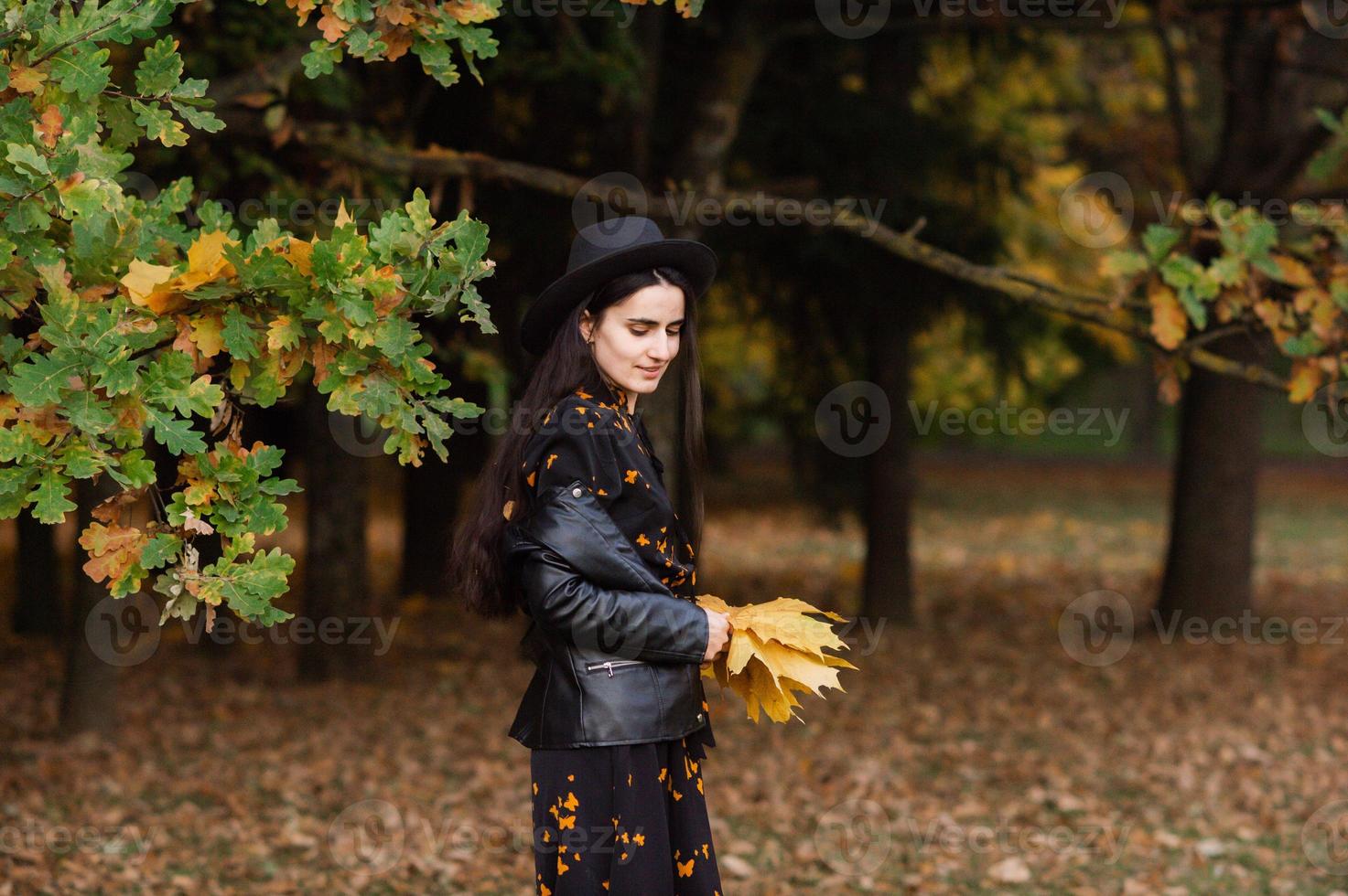 un niña en un negro sombrero sostiene amarillo otoño hojas en su manos foto