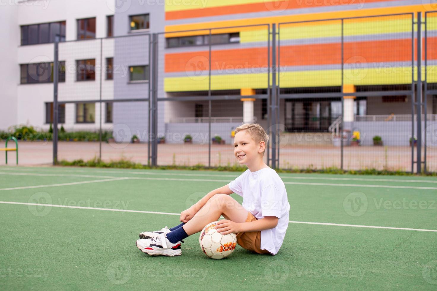 A teenage boy is sitting on a green field in the school yard with a soccer ball photo