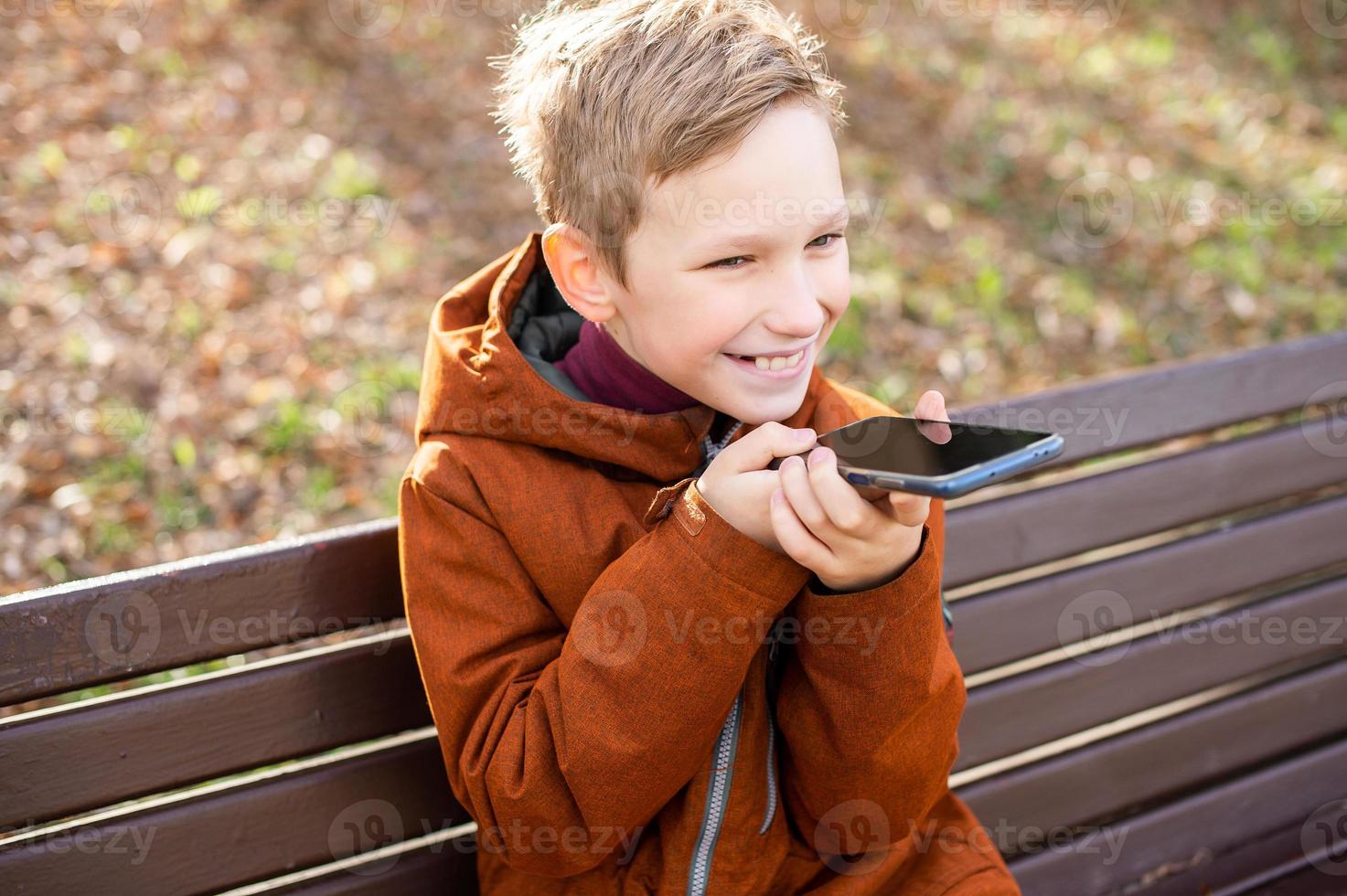 A boy records a voice message and laughs while sitting on a park bench in autumn photo