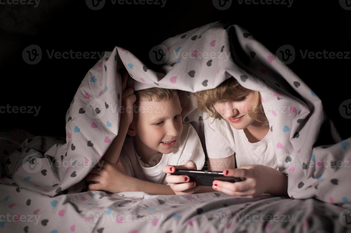 Brother and sister lie under a blanket at night and play on the phone photo