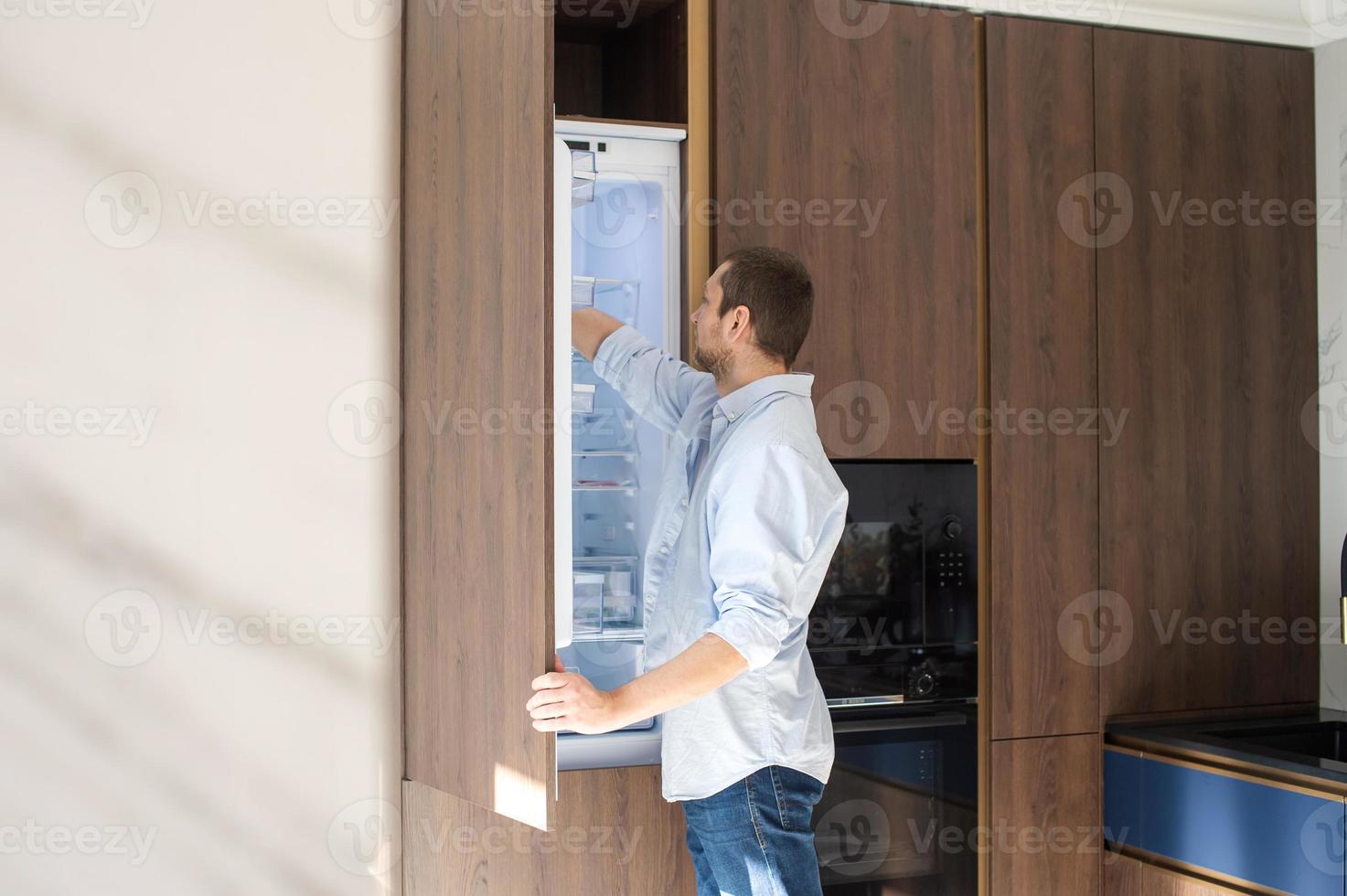 A man in a shirt opened the refrigerator and takes out groceries photo