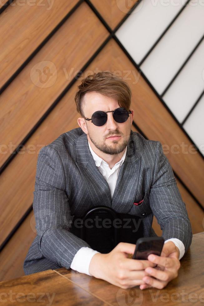 A young man in a suit and sunglasses is sitting at a desk in the office and looking at the phone photo