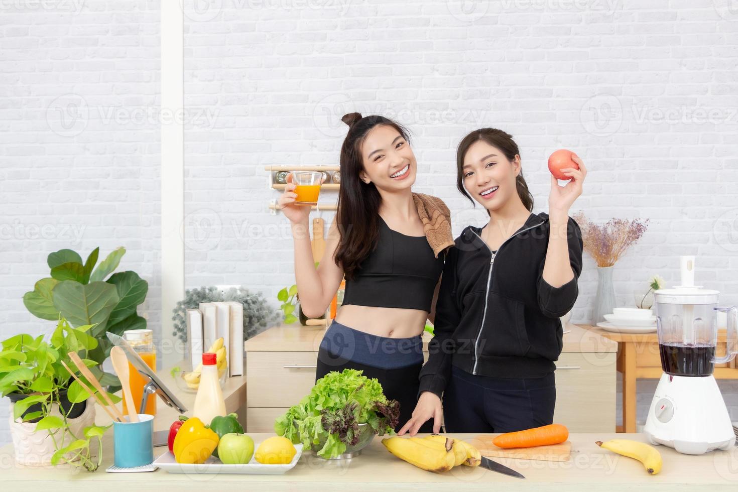 Asian active two women sibling in sportswear enjoy eating fruits healthy foods. Beautiful girl, sister holding a glass of orange juice and apples in the kitchen photo