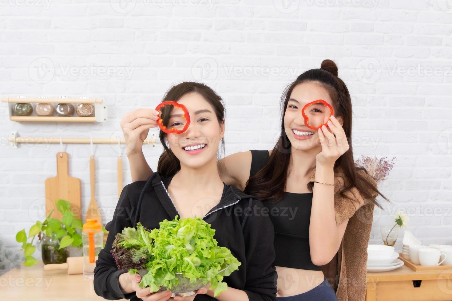 Asian active two women sibling in sportswear enjoy eating fruits healthy foods. Beautiful girl, sister holding Bell Pepper, sliced like glasses. In the kitchen. photo