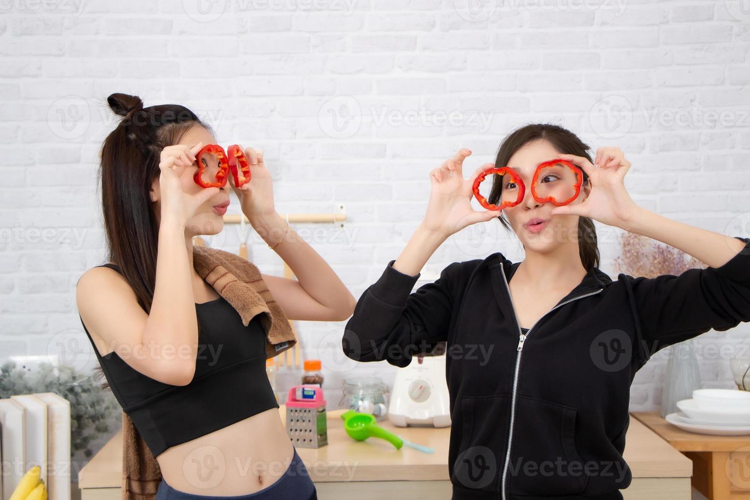 Asian active two women sibling in sportswear enjoy eating fruits healthy foods. Beautiful girl, sister holding Bell Pepper, sliced like glasses. In the kitchen. photo