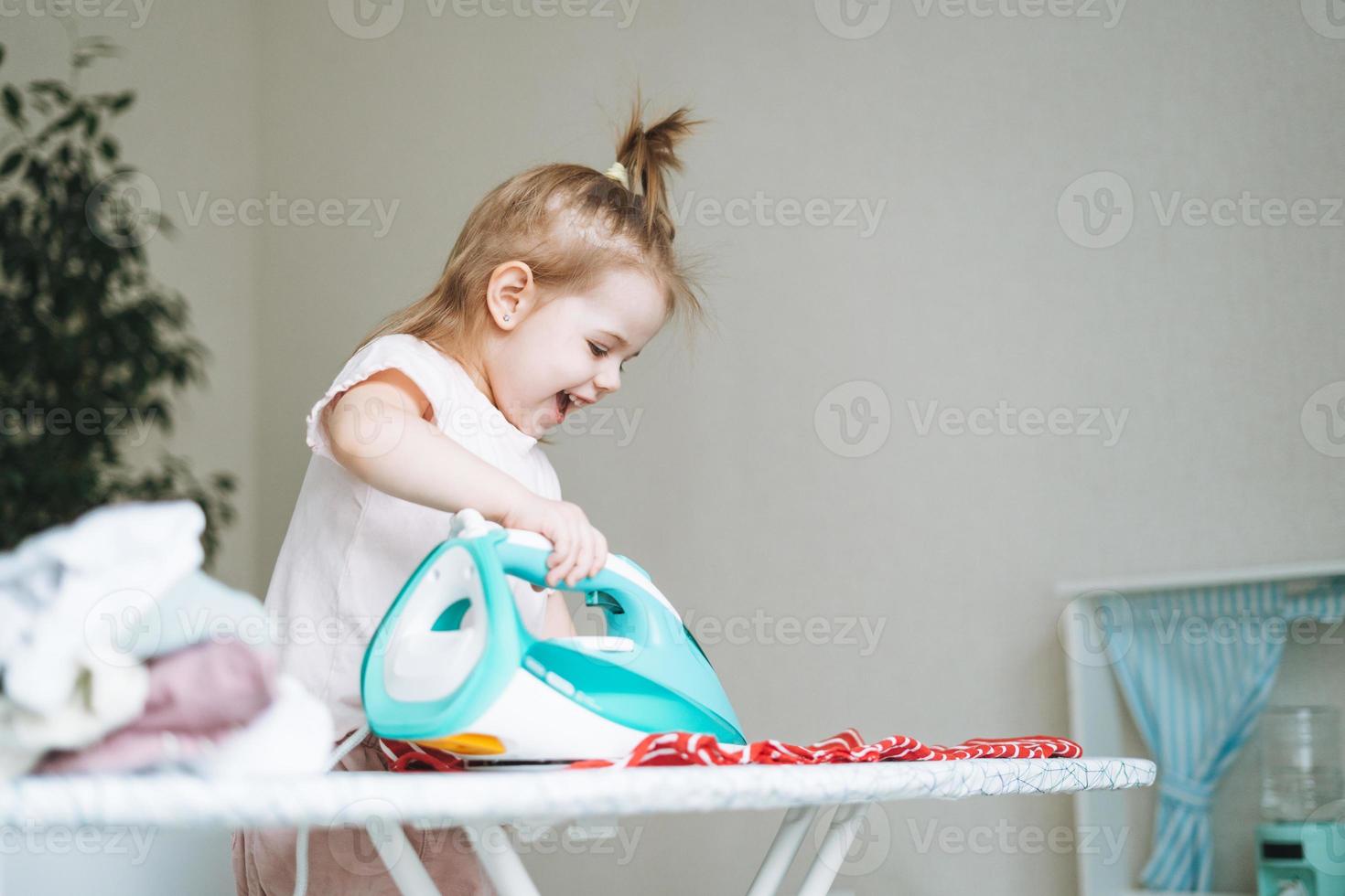 linda pequeño bebé niña jugando y planchado ropa con un hierro a hogar foto