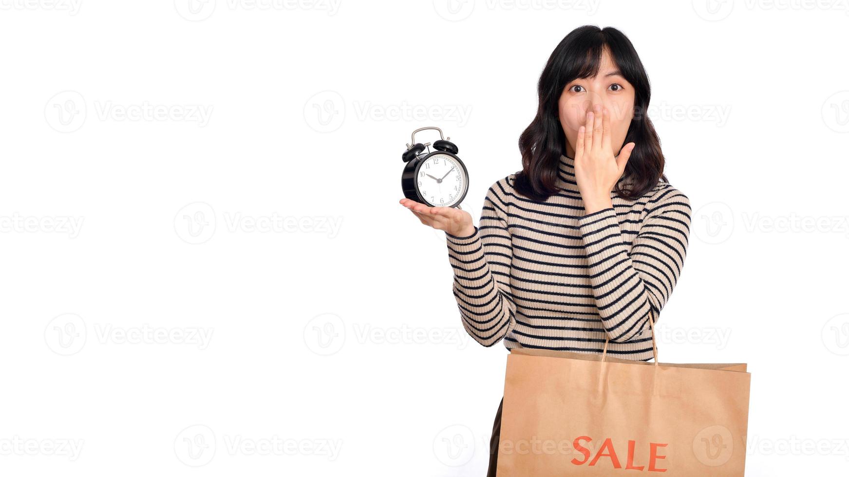 Portrait of excited young Asian woman with sweater shirt holding alarm clock and shopping paper bag isolated on white background. last deals shocked price shopping concept photo