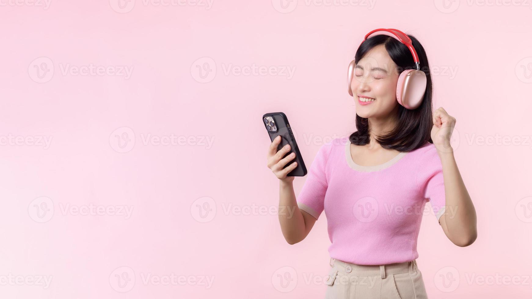 Portrait cheerful young asian woman enjoy listening audio by smartphone music application against pink. Happy smiling female person with headphone. Sound, leisure, lifestyle, technology concept photo