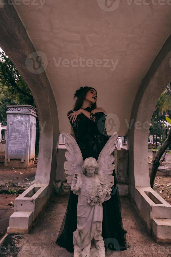 a young witch in a black dress is standing between the tombstones shaped like an angel while visiting the cemetery photo