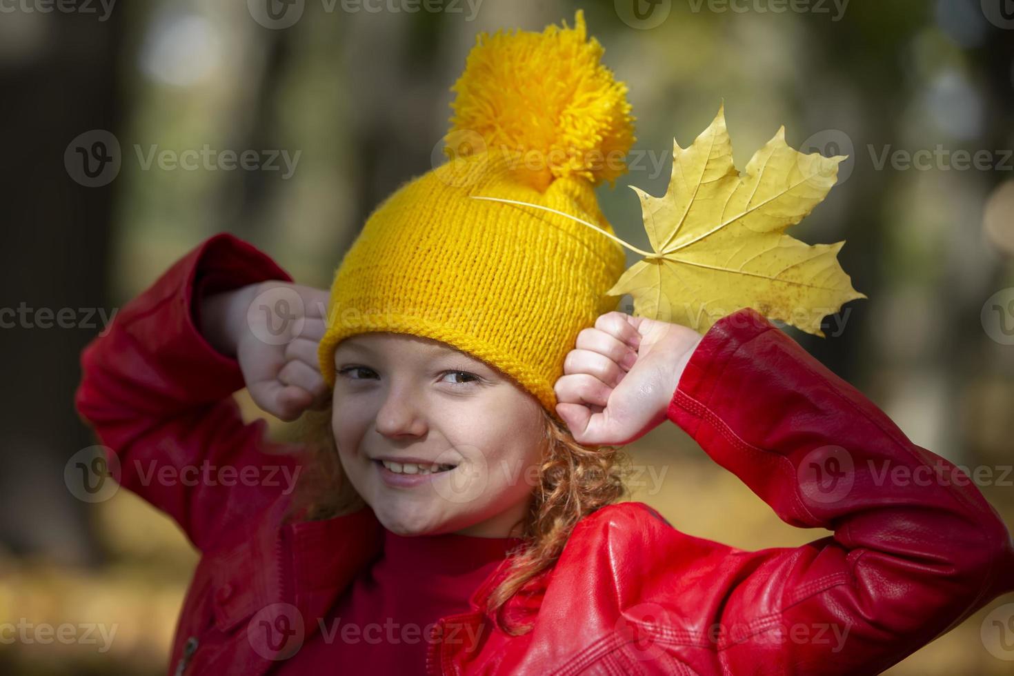 Girl in a yellow knitted hat in autumn. Portrait of a child in the autumn park. Child holding autumn maple leaves. photo