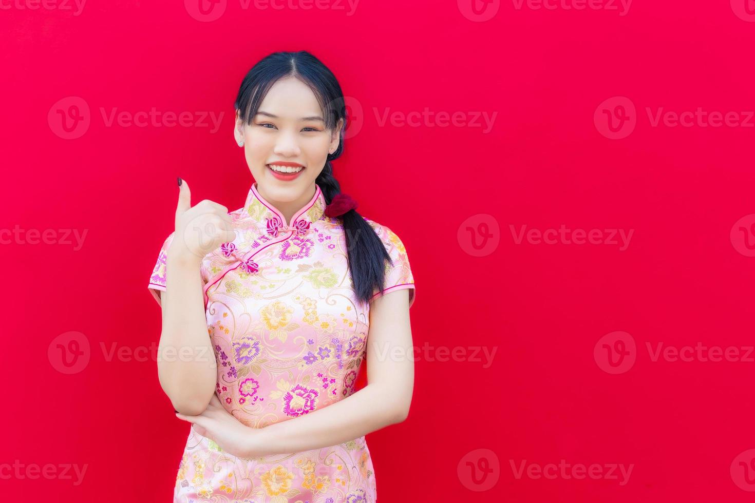 Beautiful Asian woman wears pink cheongsam and arm crossed while looks at to camera and smiles happily and acting hand shows thumbs up  with the red background,Celebrate Chinese New Year Theme. photo