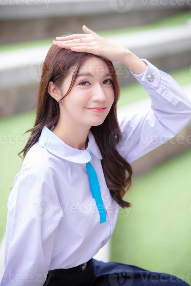 Beautiful high school Asian student girl in the school uniform sitting and smiles happily while she put her hand on head confidently  with the building as a background. photo