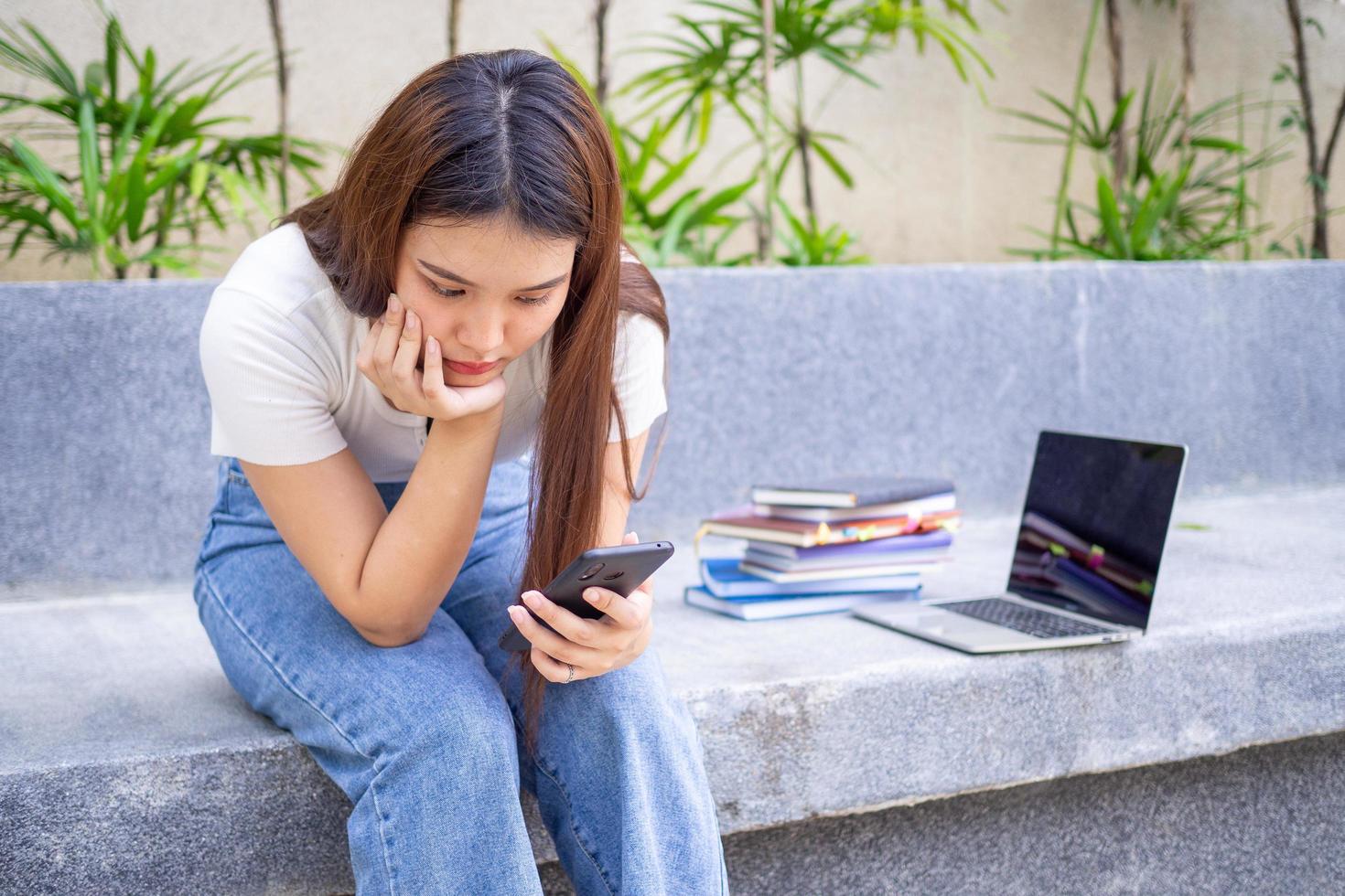 un asiático hembra estudiante es sentado en el jardín siguiente a el teléfono, mirando a el resultados de un en línea prueba en un teléfono inteligente, en el cara de decepción y lamentar terminado el información en el teléfono foto