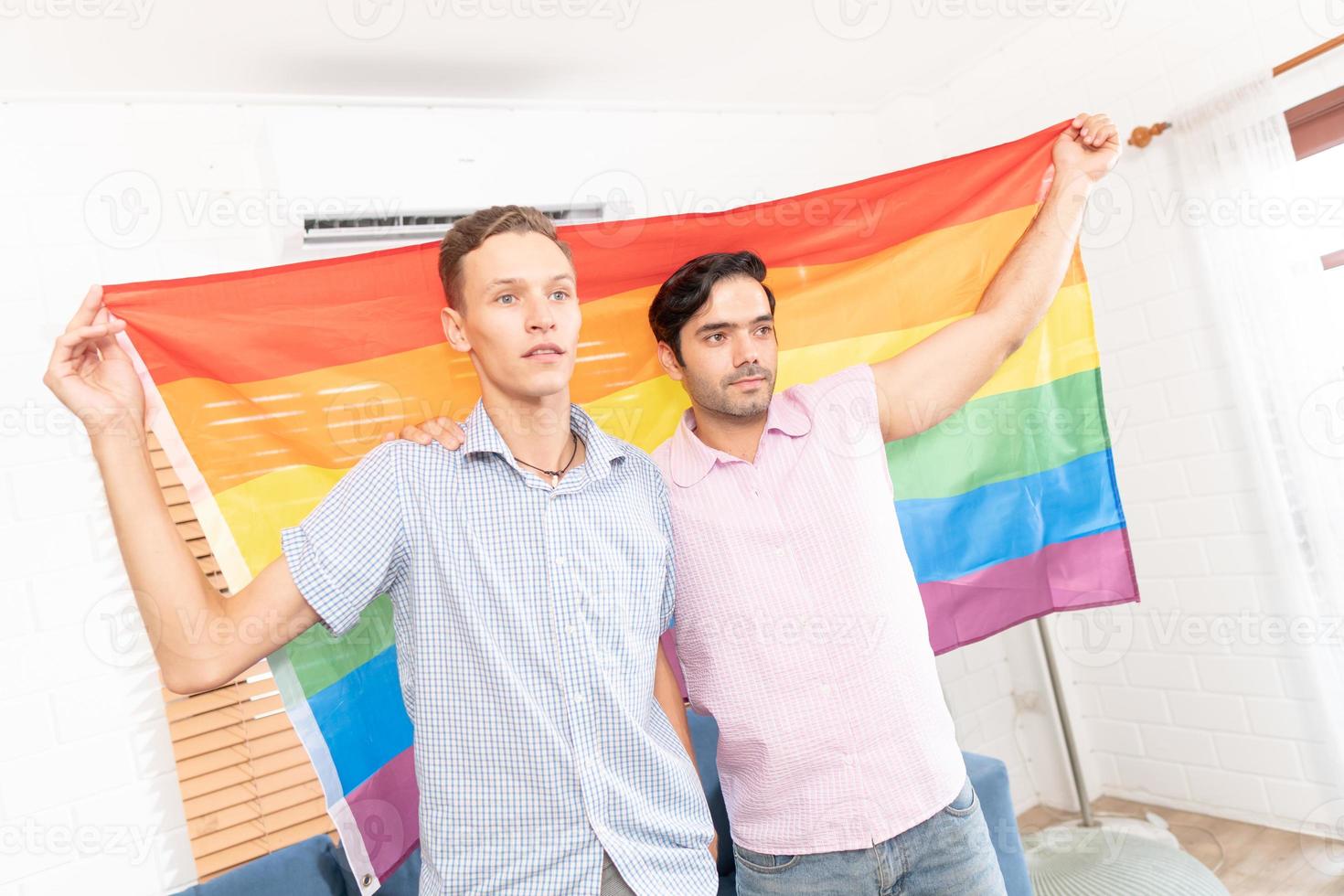Happy LGBT couple, sitting on the sofa holding and waving rainbow LGBT Pride flag together in the bedroom at home. Diversity of LGBT relationships. A gay couple concept. Loving gay couple. photo