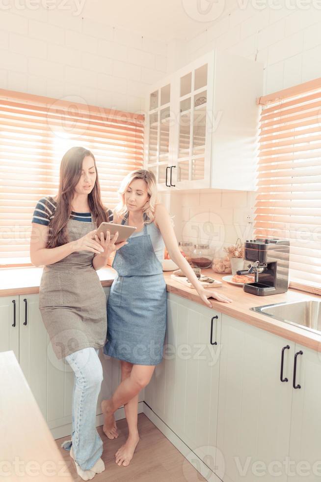 hermosa lesbiana Pareja Cocinando sano alimento, mirando en digital tableta a moderno cocina interior. homosexual relaciones y sano comiendo concepto. lgbt, lgbtq hogar en línea cocina. calentar tono. foto