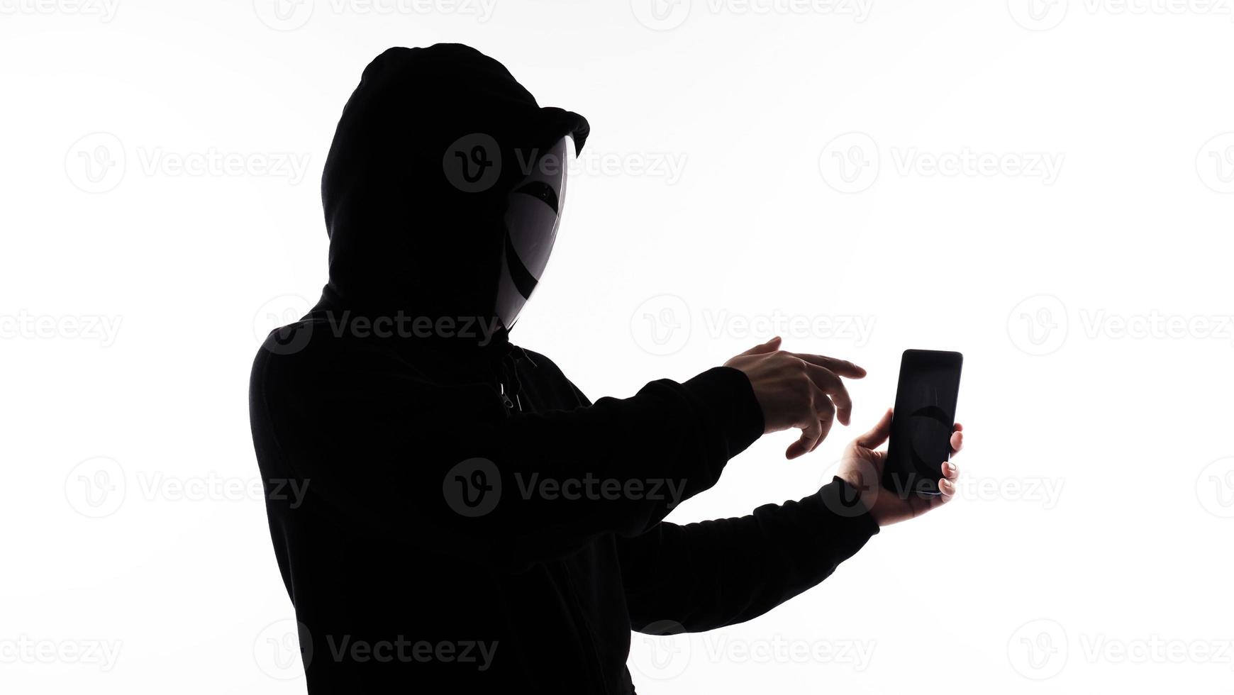 hacker anónimo y mascarilla con smartphone en la mano. hombre con camisa de capucha negra sosteniendo y usando un teléfono móvil con fondo blanco. representan el concepto de piratería informática o robo de datos personales foto