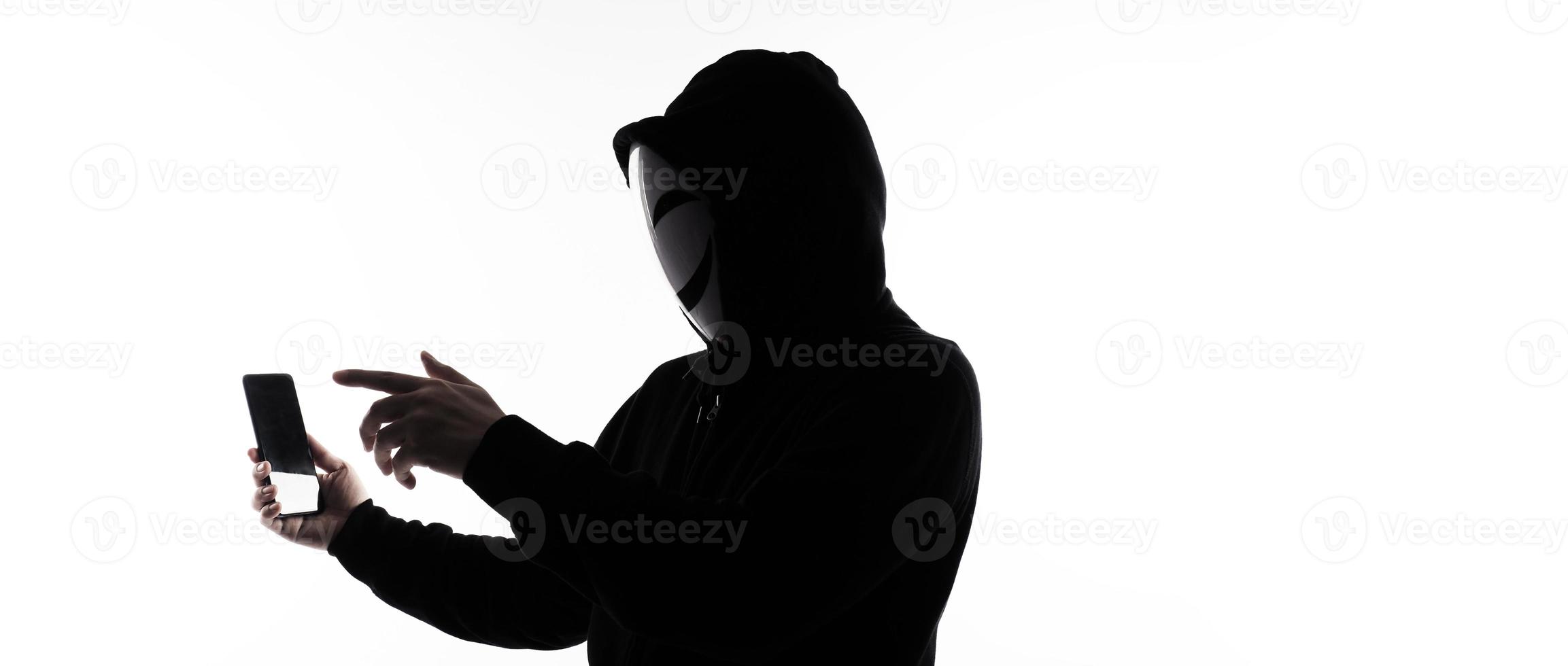 Hacker Anonymous and face mask with smartphone in hand. Man in black hood shirt holding and using mobile phone on white background. Represent cyber crime data hacking or stealing personal data concept photo