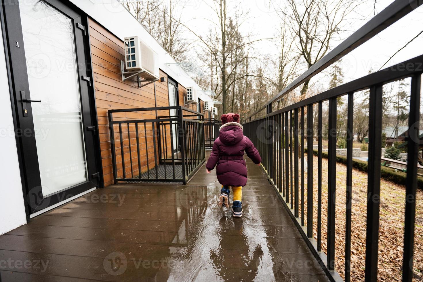 Back of baby girl walking on terrace of one-storey modular houses in spring rainy forest. photo