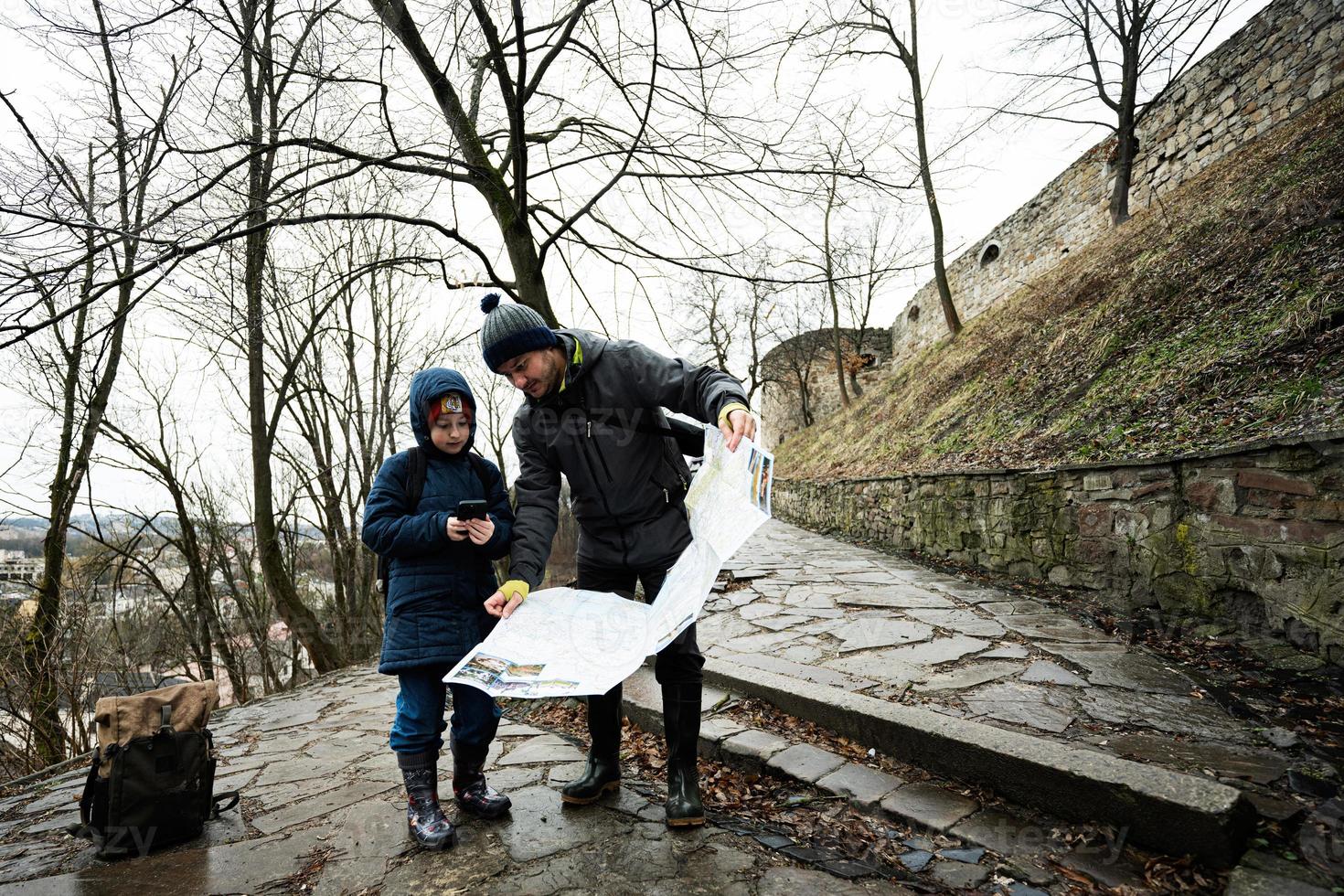 Father and son tourist look at map, stand on wet path to an ancient medieval castle fortress in rain. photo