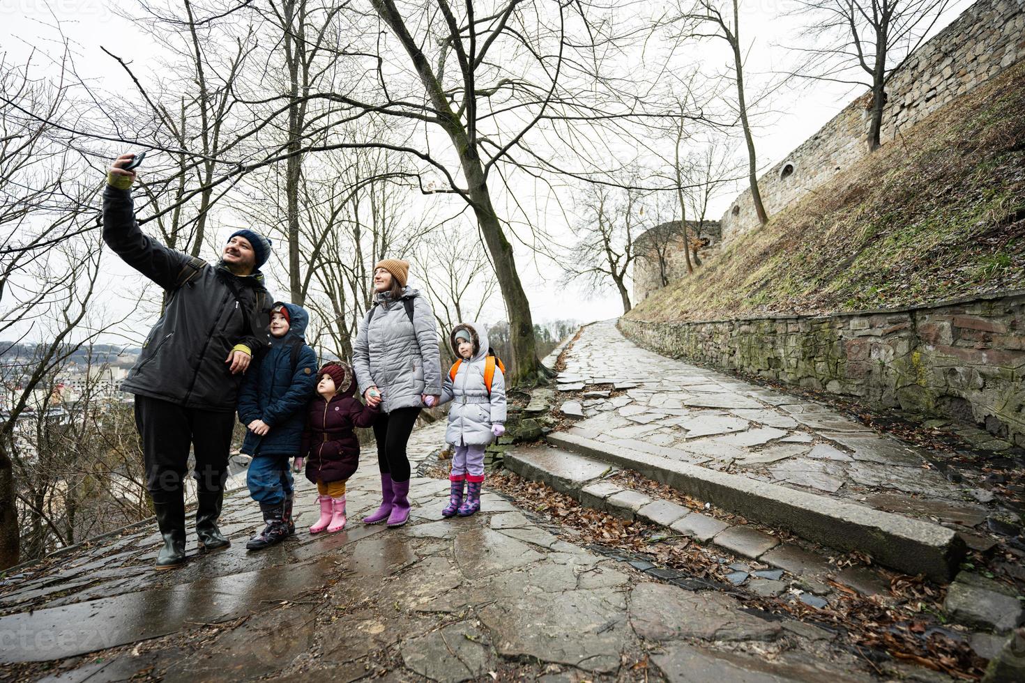Family tourist with three children stand on wet path to an ancient medieval castle fortress in rain and making selfie photo on phone.