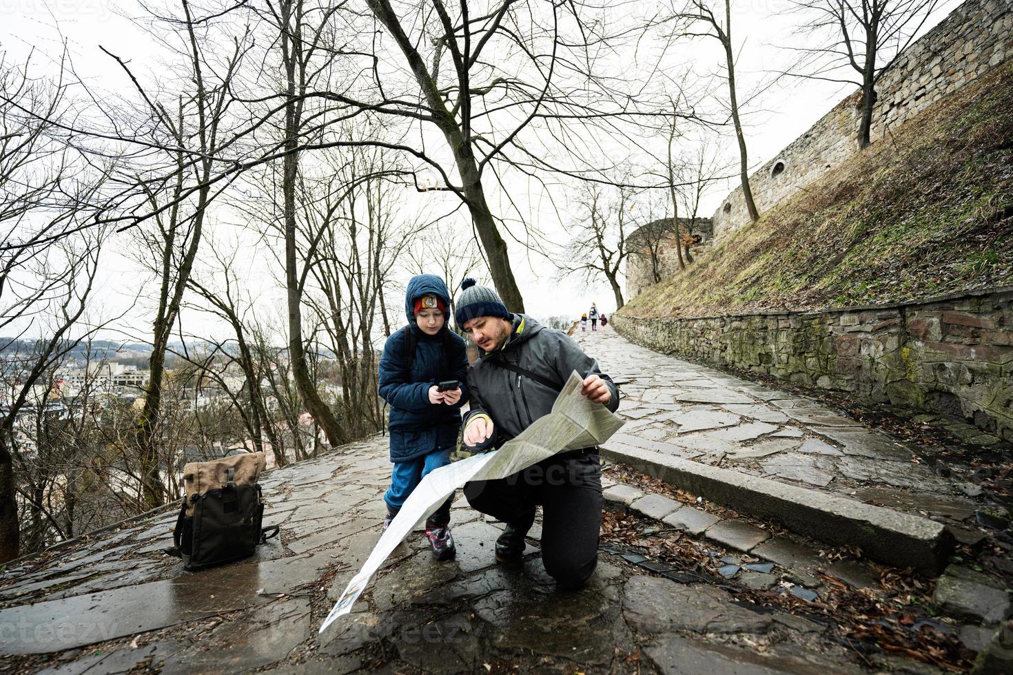 Father and son tourist look at map, stand on wet path to an ancient medieval castle fortress in rain. photo