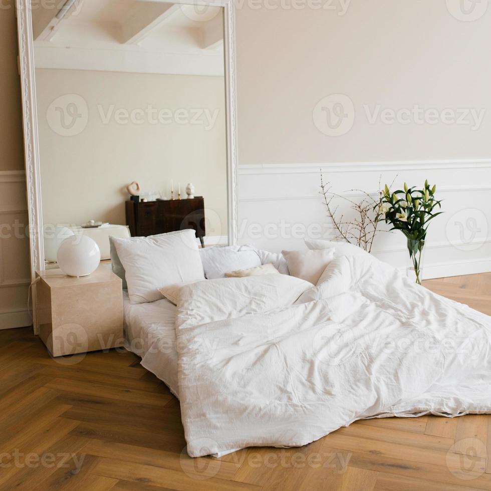 A double bed with white linens and beige pillows in a bright Scandinavian bedroom, a bed on the floor and flowers in a vase, a large mirror in a frame photo