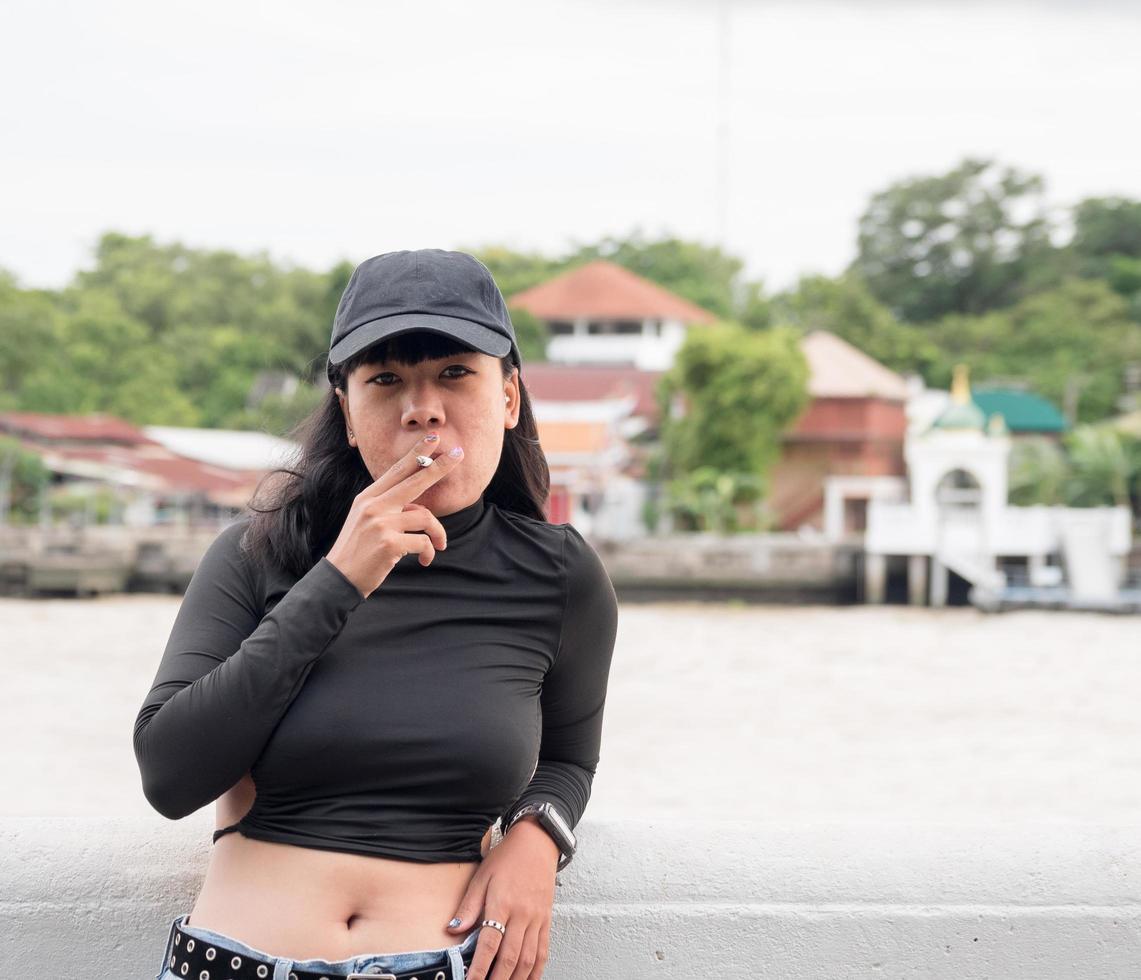 retrato mujer niña adolescente joven Asia uno persona vistiendo un sombrero y largo negro pelo vistiendo un negro camisa mano sostener de fumar cigarrillo blanco color en pie al aire libre por el pared al aire libre foto
