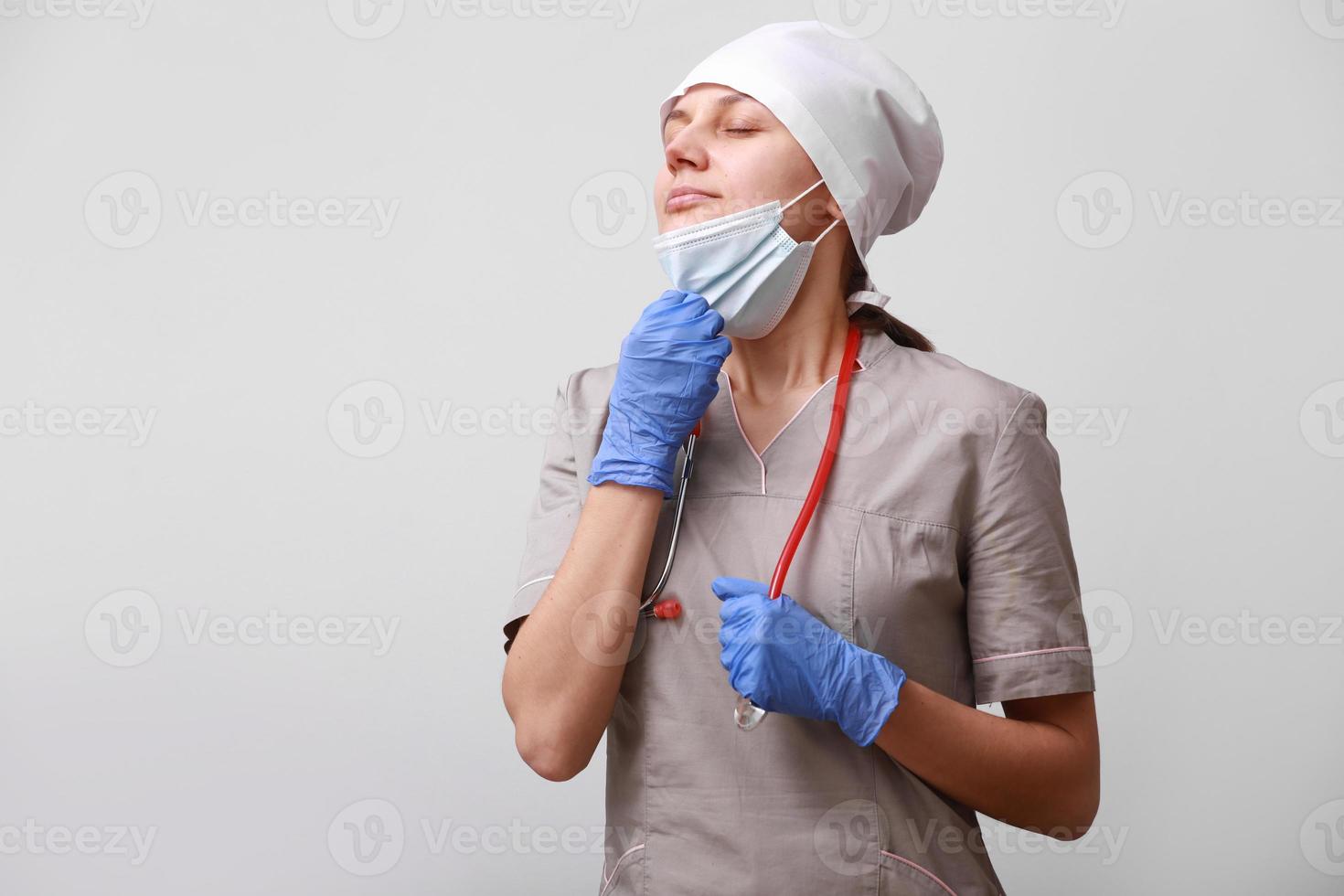 smiling female doctor with stethoscope in uniform. Woman is taking off medical protective mask from face to breathe air, on white background. Difficulties and problems at work of a medical worker. photo
