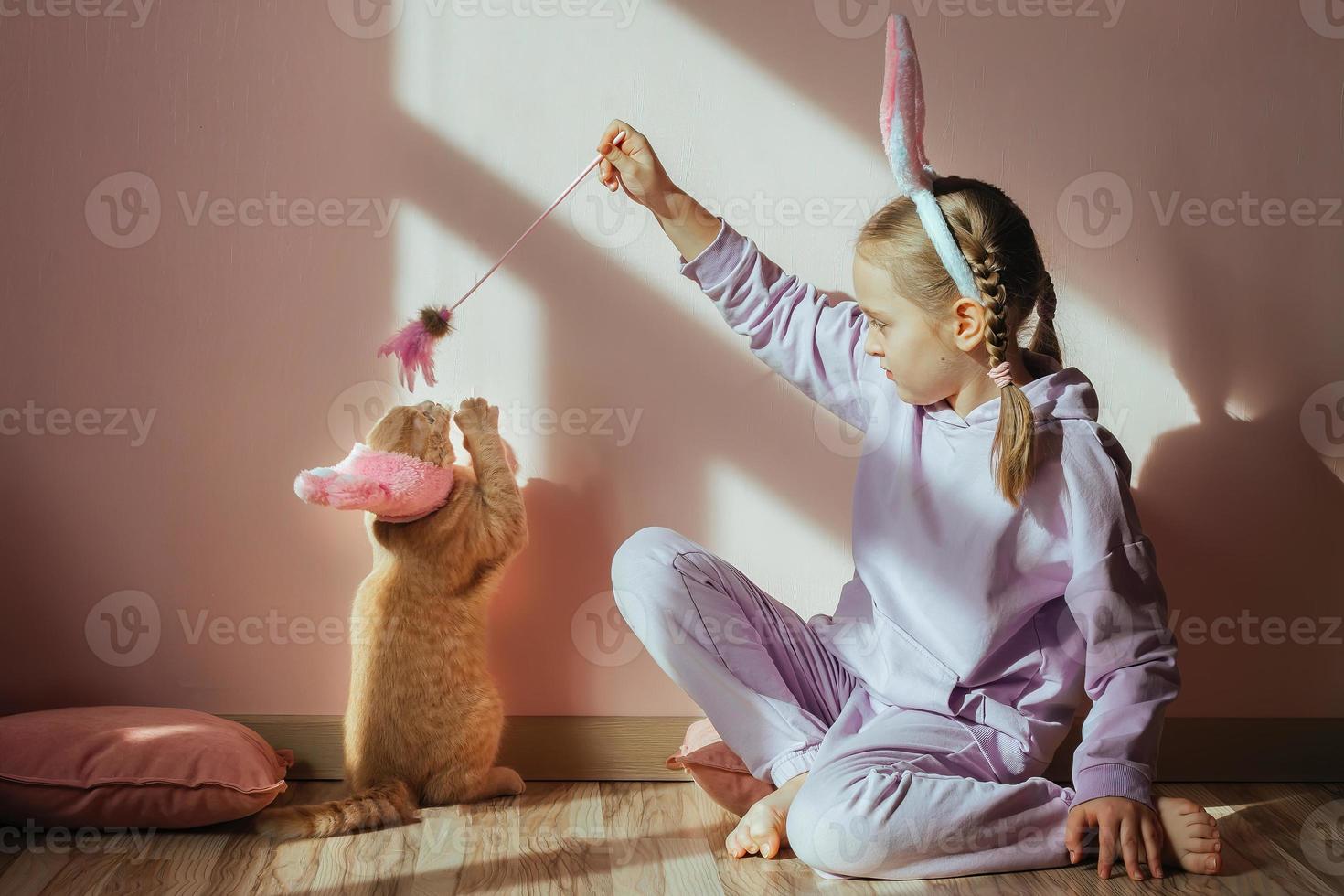 una colegiala en la cabeza una diadema con orejas de conejo juega con un gato doméstico rojo en un sombrero de pascua al sol foto
