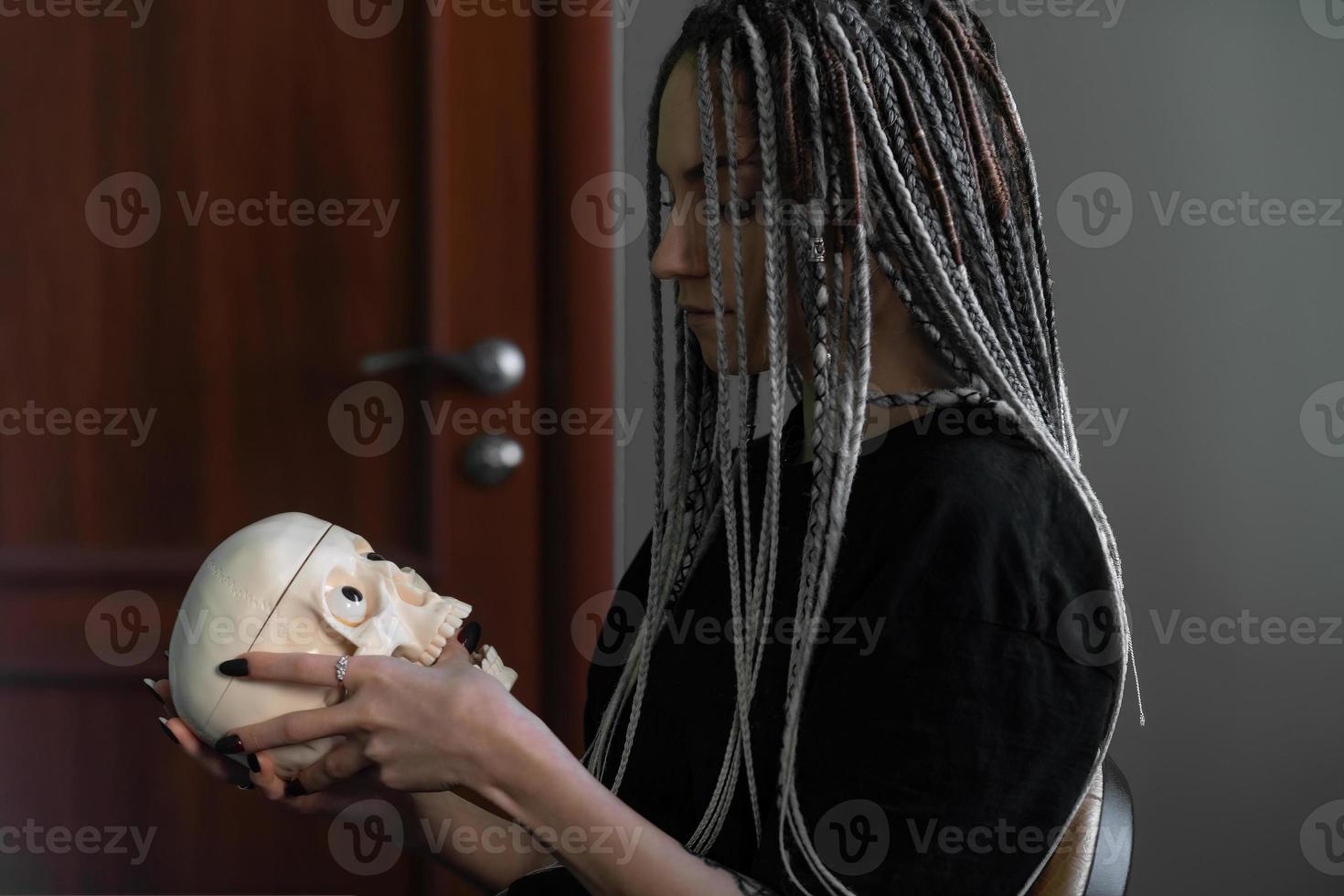 A young bright girl in dark clothes, with dreadlocks on her head, holds a skull in her hands. Generation z, gothic style photo
