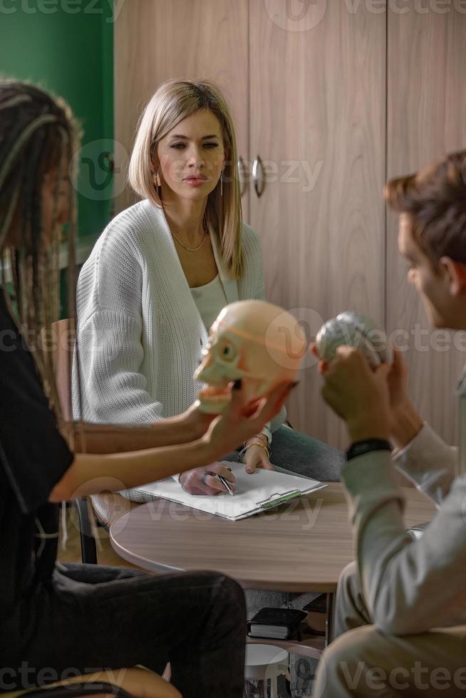 A female psychologist works in her office with a young couple. In the hands of a specialist, the notes that she makes during a consultation, young people hold a model of a human skull in their hands. photo