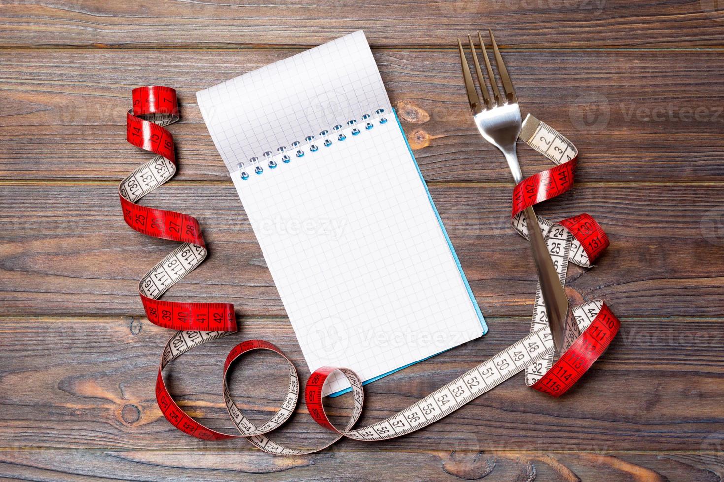 Top view of weight loss concept on dark wooden background. Mix of red centimeter tape, note pad and fork with space for your design photo