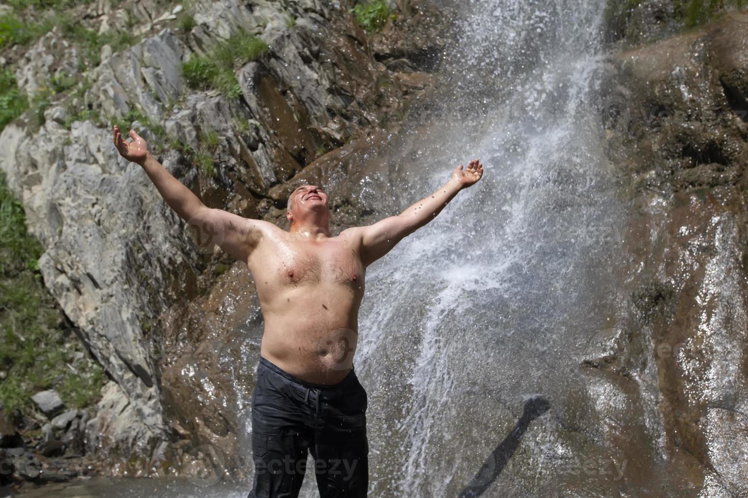 An elderly man stands at a mountain waterfall, spread his arms and enjoys the moment. photo