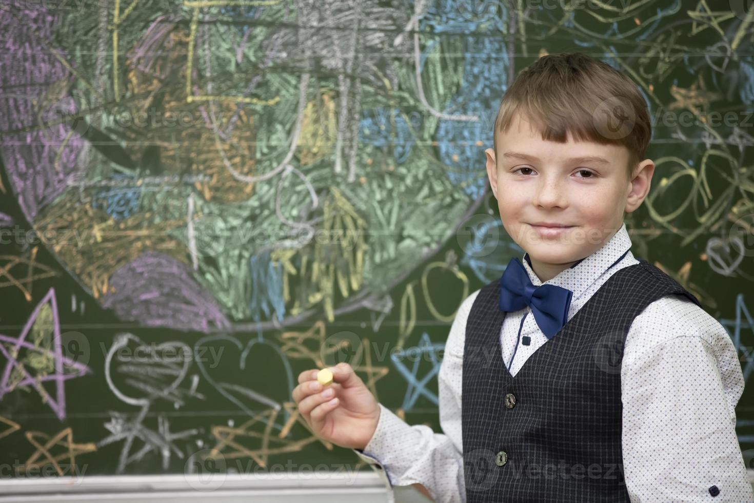 A handsome boy of school age stands at the blackboard, looks at the camera and smiles. Smart student. Back to school. photo