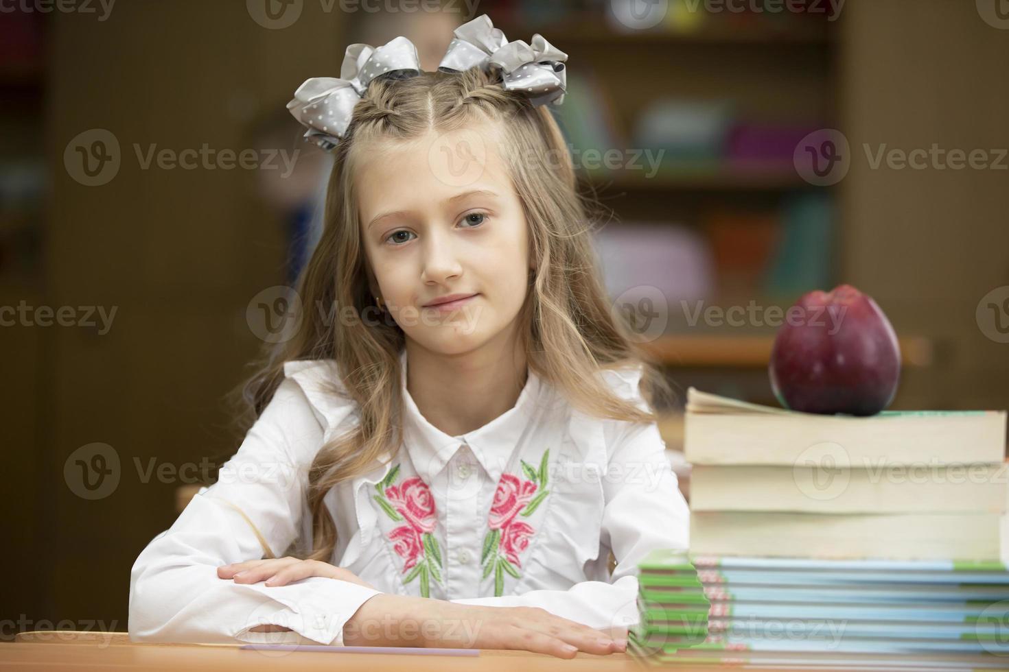 Beautiful schoolgirl with bows at her desk. Girl in the classroom with books and an apple. Secondary school. Back to school. photo