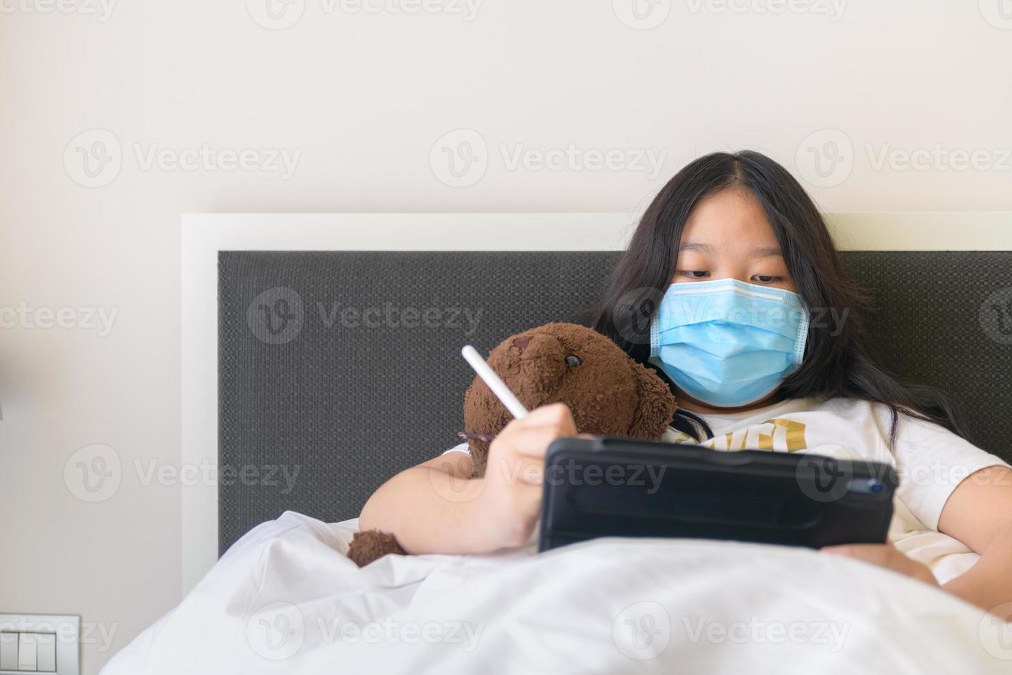 enfermo niña vistiendo máscara y estudiando en línea con tableta pn cama, nuevo normal foto