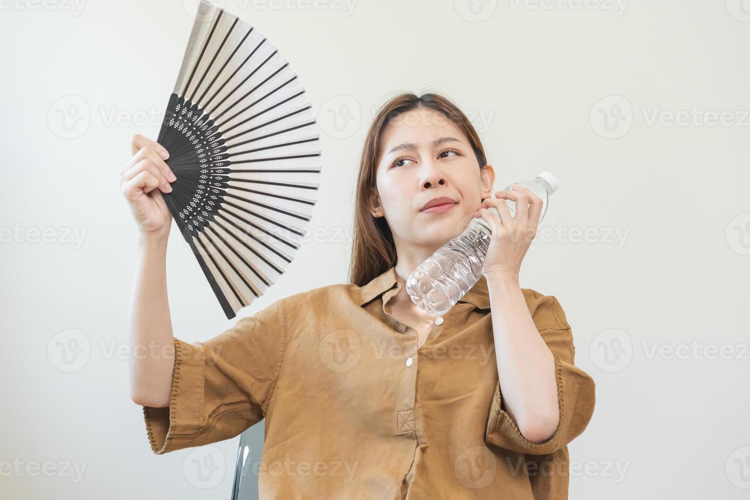 Summer heat stroke, hot weather, tired asian young woman sweaty and thirsty, refreshing with hand in blowing, wave fan to ventilation, holding cold water bottle tap her body when temperature high. photo