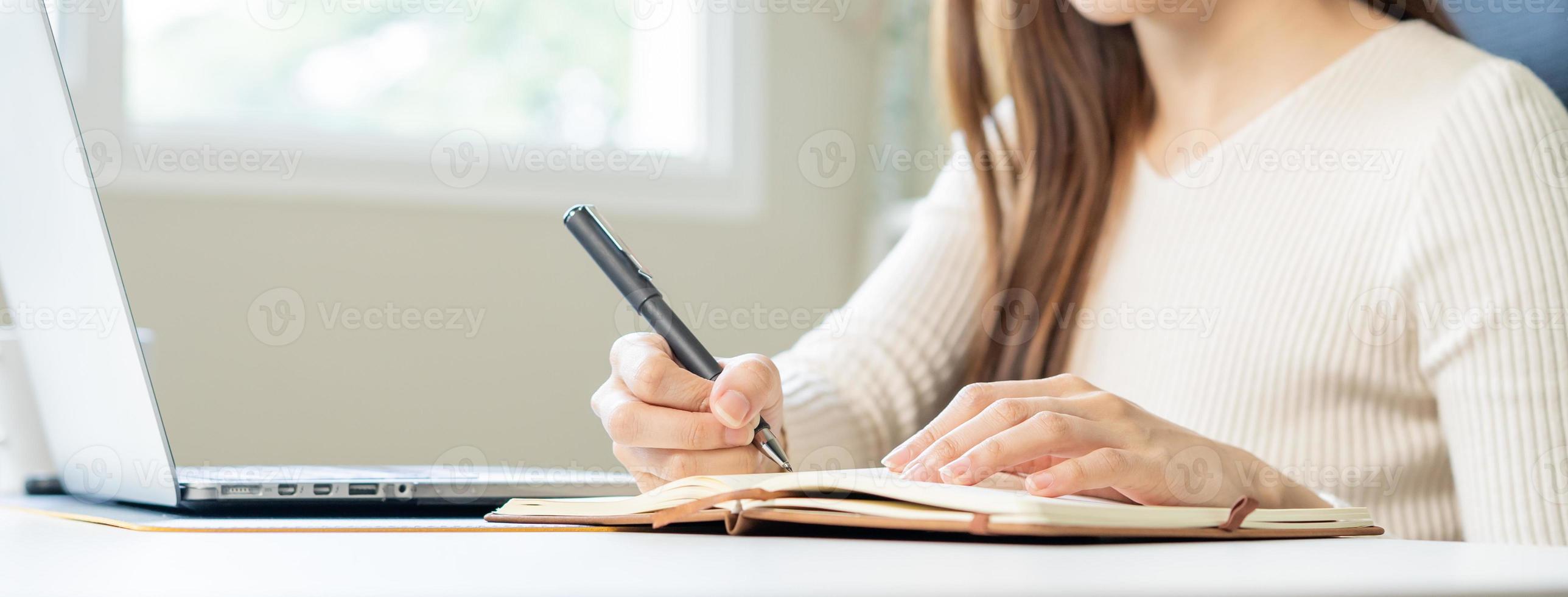 Video conference, asian young woman working, hand in make notes on notebook, using laptop computer for online meeting in video call, learning education, writing while work sitting, distance from home. photo