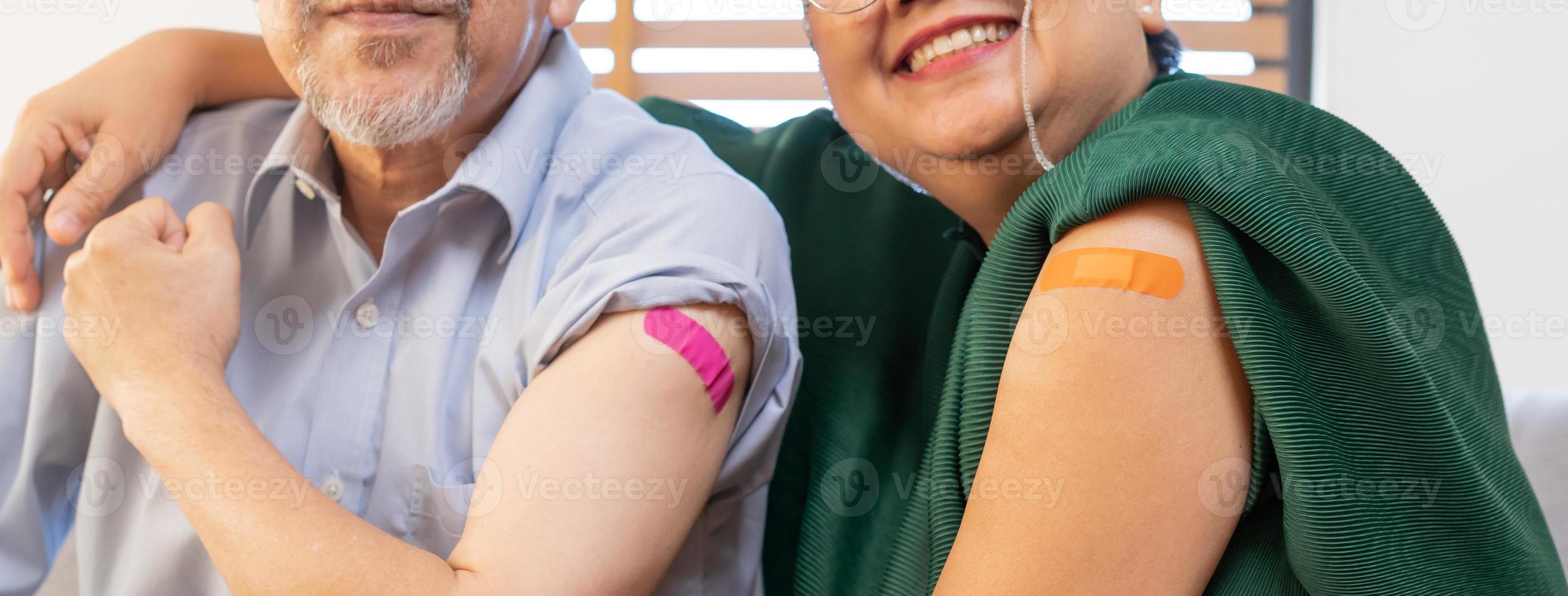 Showing bandage on arm, shoulder, happy asian elderly, aged family smile strong together with protect of covid-19, coronavirus after injection of vaccine, sitting on couch in living room at home. photo