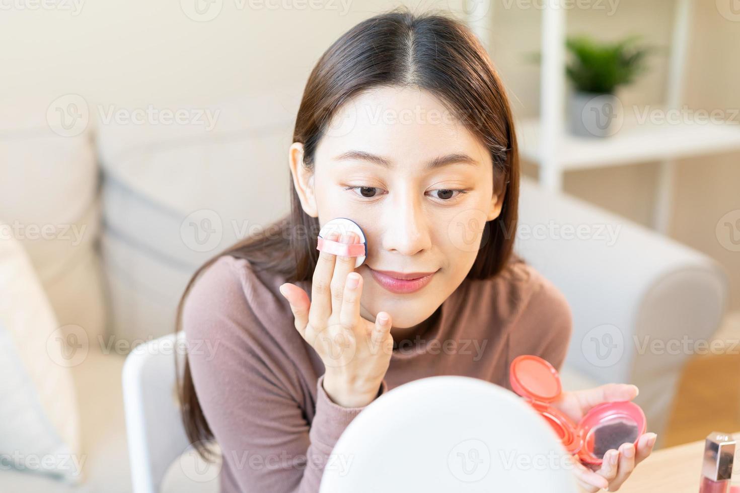 contento belleza blogger concepto, bonito asiático joven mujer, niña hacer arriba cara por aplicando rosado sonrojo por cepillo en mejilla, mirando a el espejo en mesa a hogar. hembra Mira con natural Moda estilo. foto