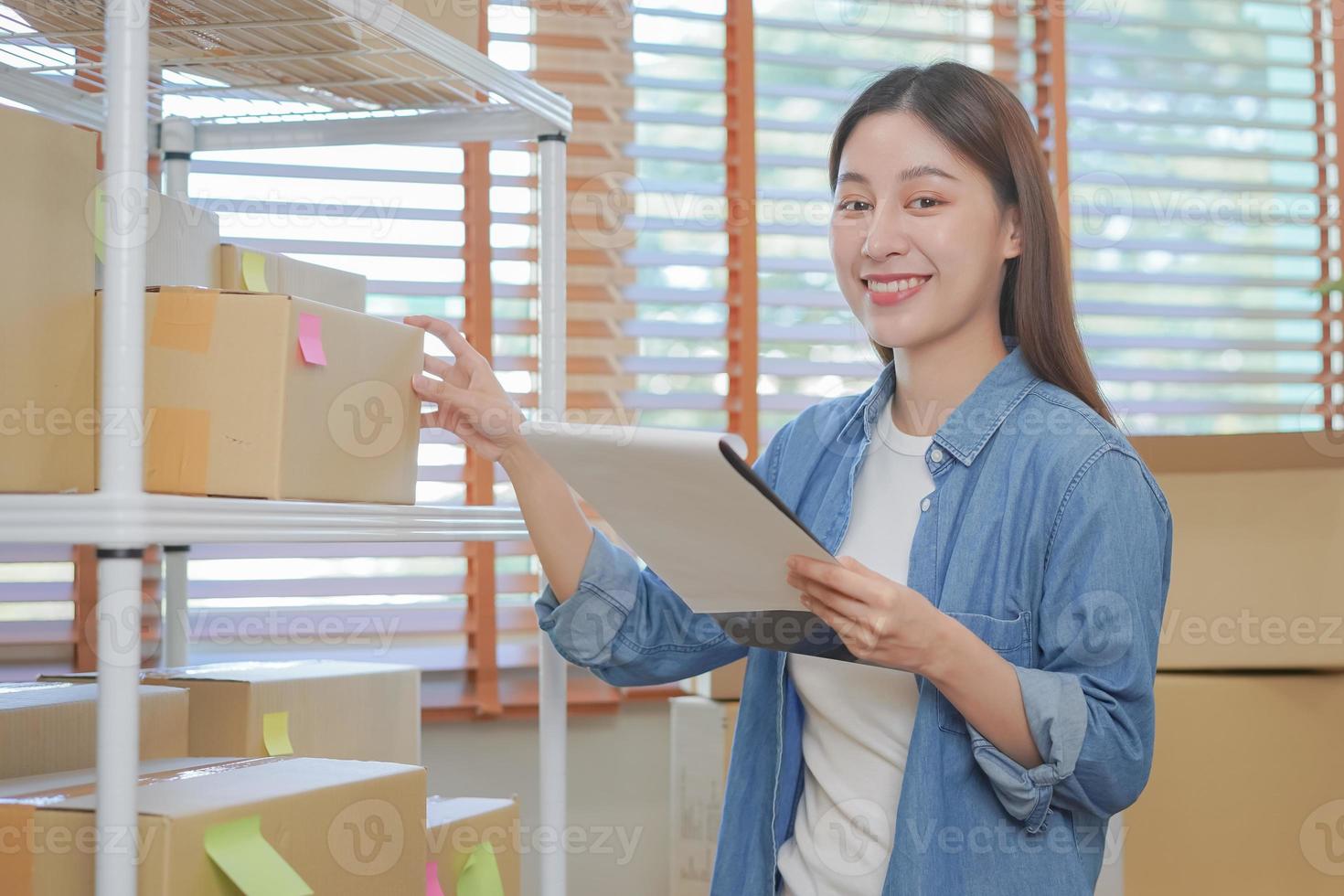 pequeño negocio empresario pequeño, sonrisa asiático joven mujer, niña propietario participación portapapeles para cheque valores de producto en estante, embalaje paquete o empaquetar a entrega después recibido orden. comerciante en línea, comercio electrónico foto