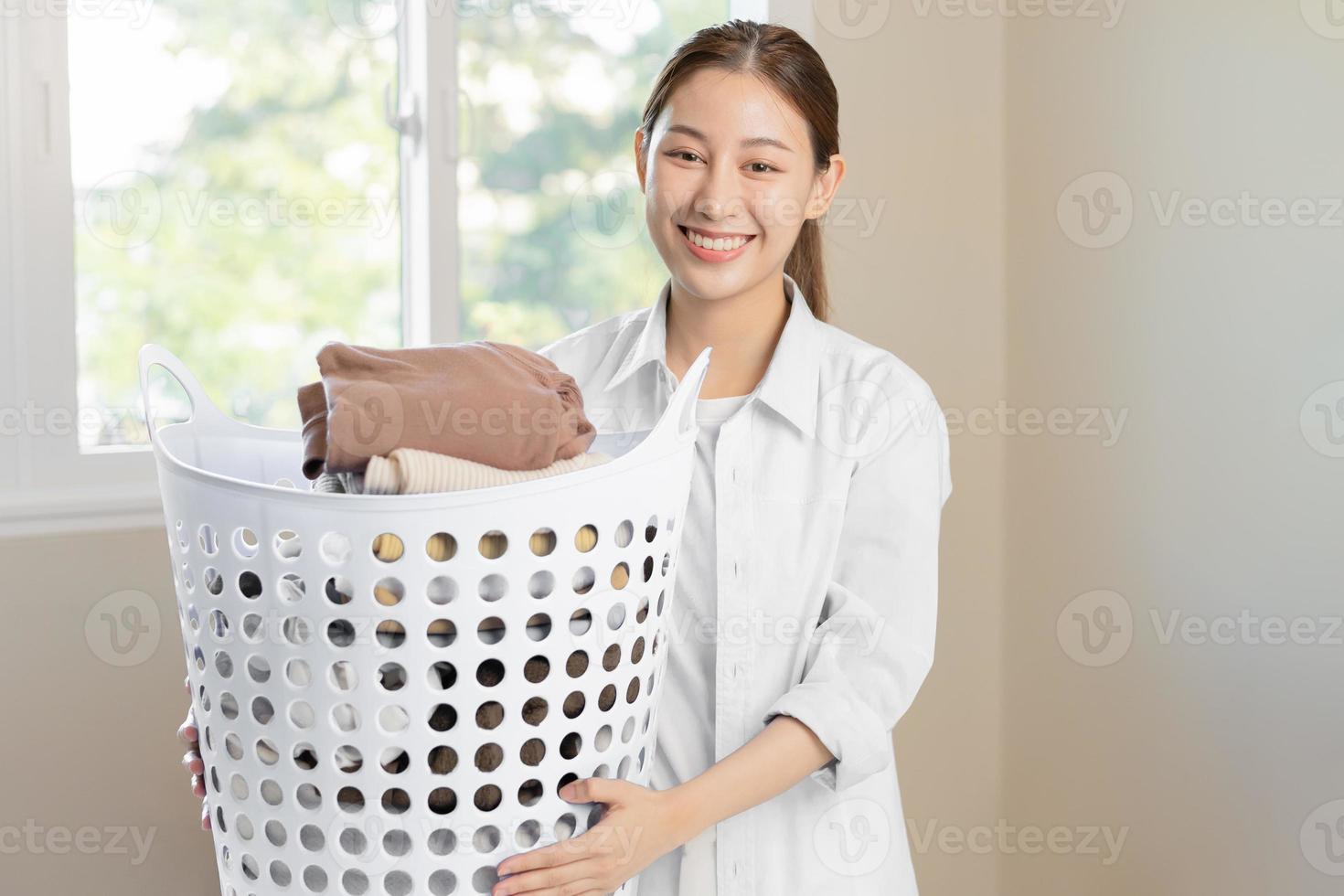 Housewife, attractive asian young woman, girl holding many folding clothes in the basket after washing clean clothes and drying, Household working in room at home, maid chore laundry tidy. photo