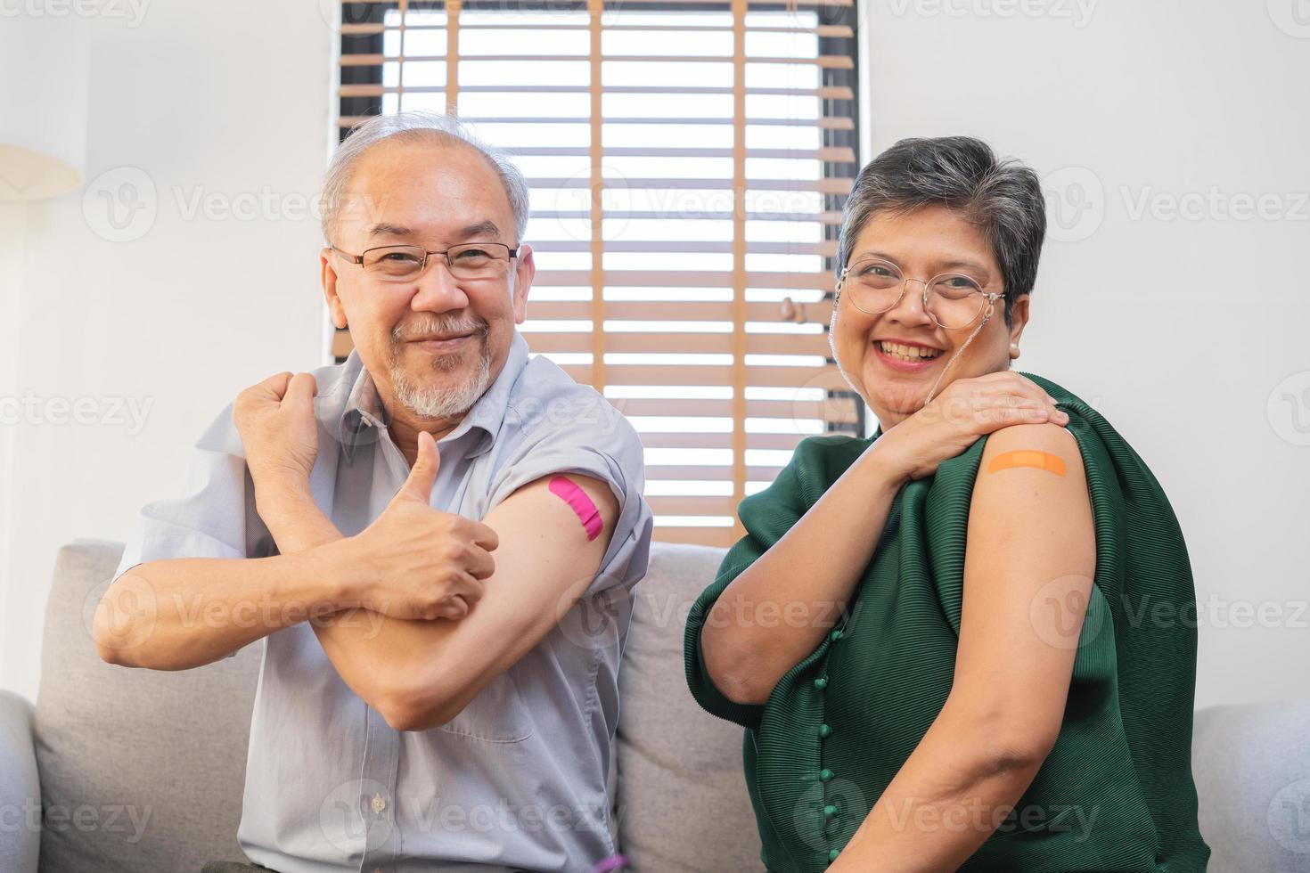 coronavirus vacunación, contento asiático anciano, Envejecido familia sonrisa fuerte juntos, demostración vendaje en brazo con proteger de covid-19 después inyección de vacuna, sentado en sofá en vivo habitación a hogar. foto